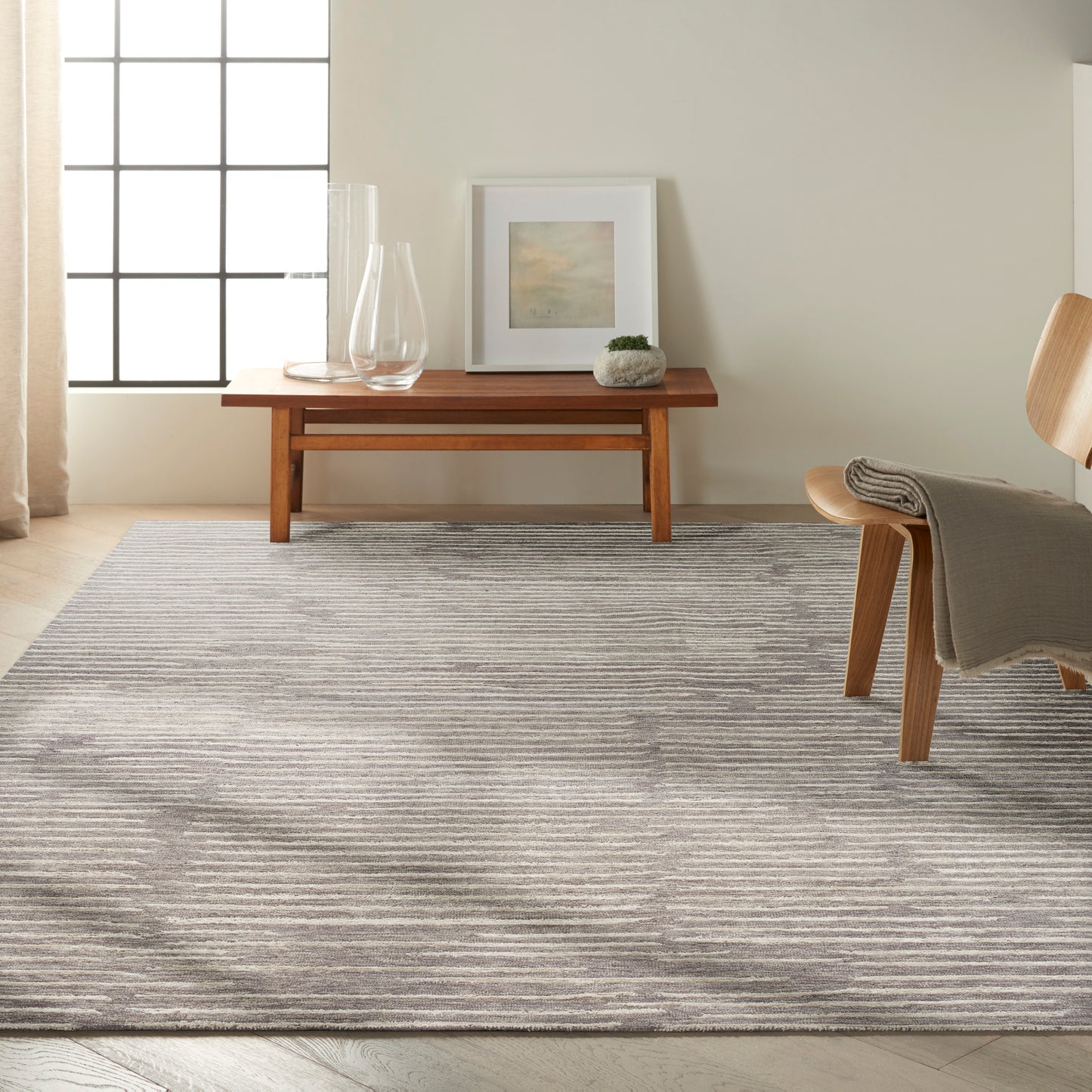 Cozy living room: light gray striped rug, wooden coffee table, glass vases, art, plant, and a wooden chair with a throw.