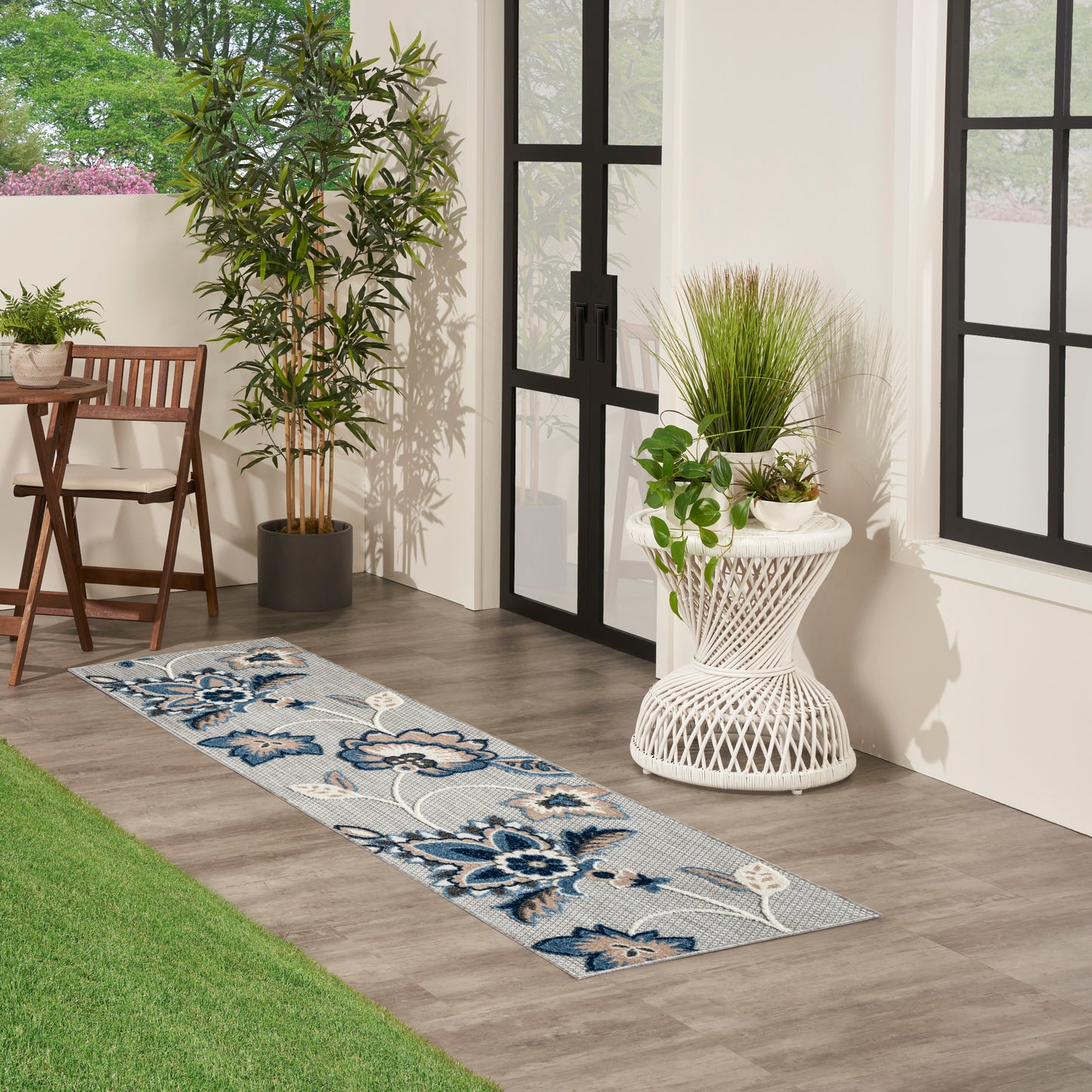 Blue floral woven runner rug on wood floor beside glass doors, with potted plants and a white wicker plant stand.