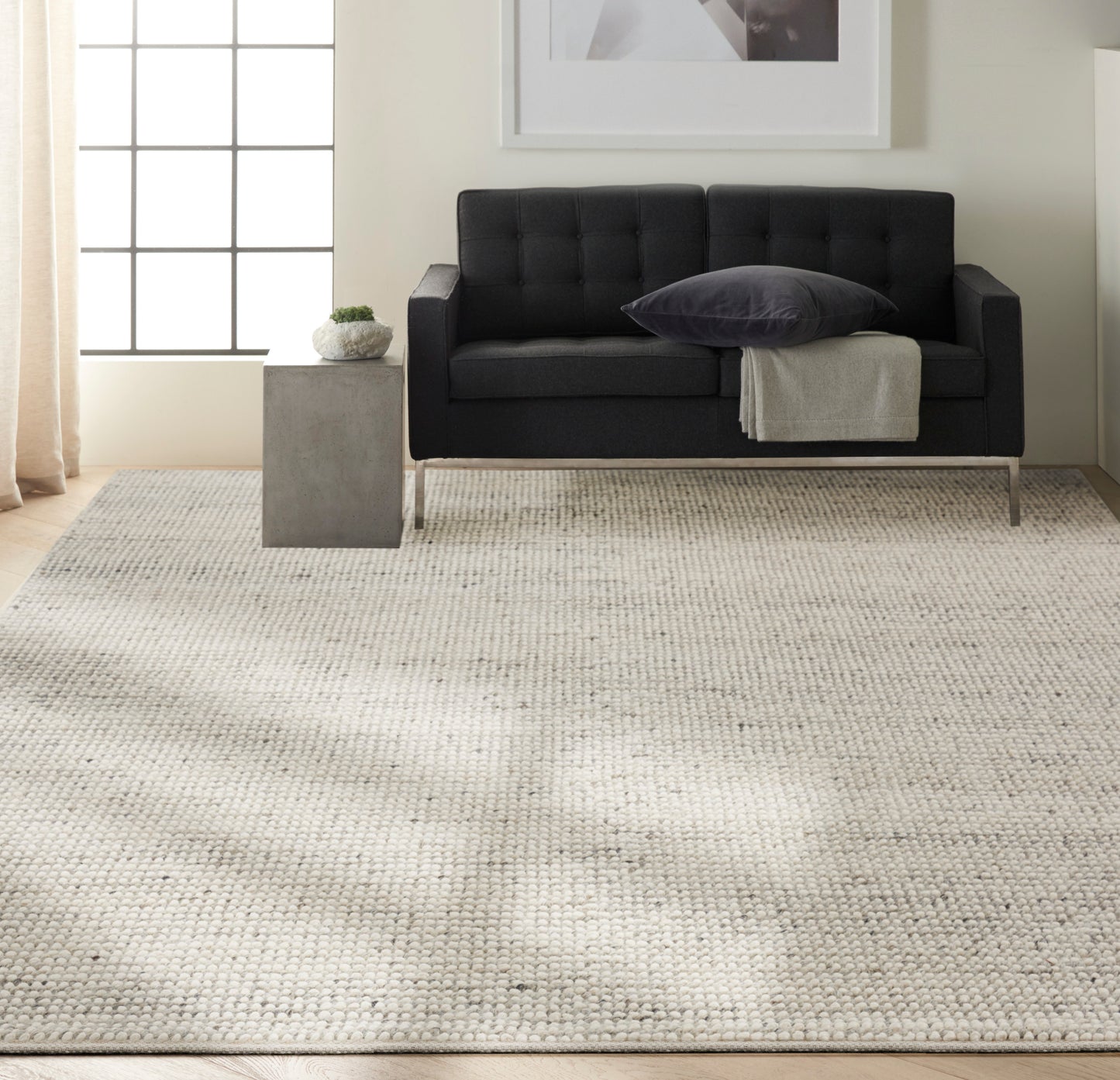 Beige woven rug in a living room scene with a dark tufted sofa, gray pillow, light throw, and a concrete side table with a plant.