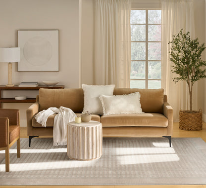 Tan sofa with white pillows and cream throw, round wooden coffee table, neutral living room with rug, plant & curtains.