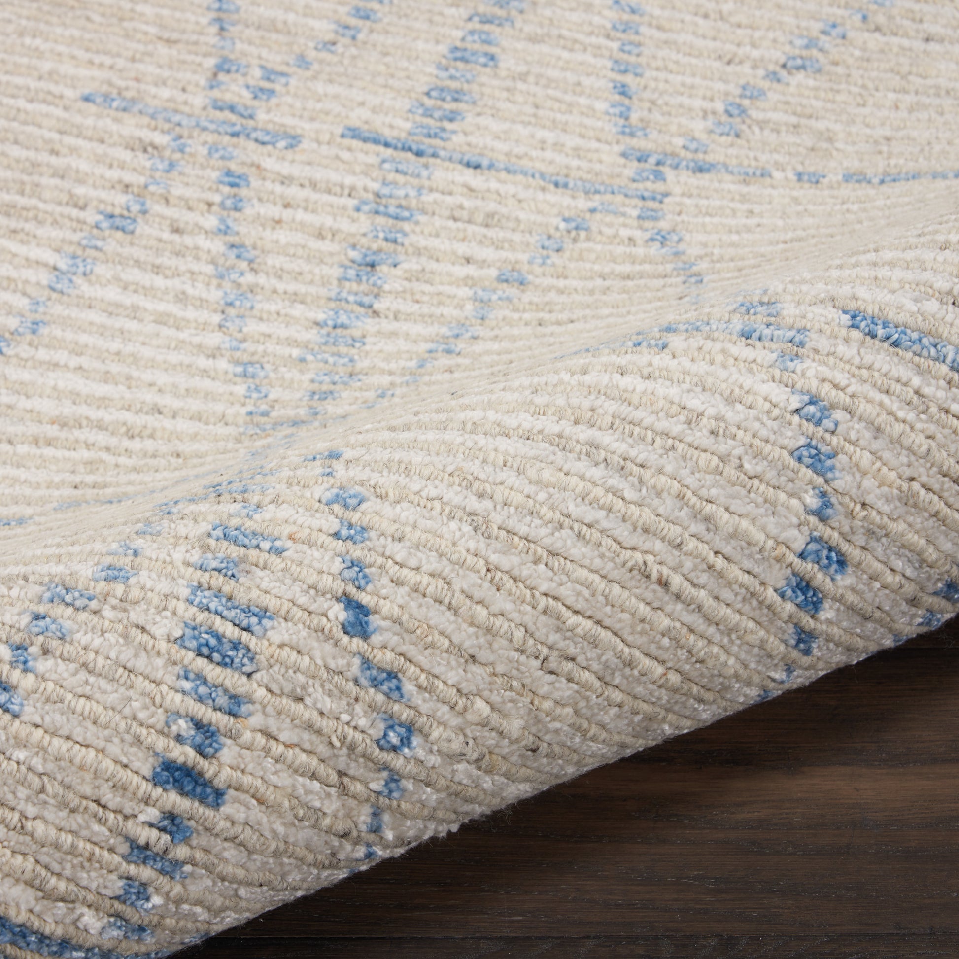 Beige textured rug with light blue geometric lines, thick woven pile, rolled edge on dark wood floor.