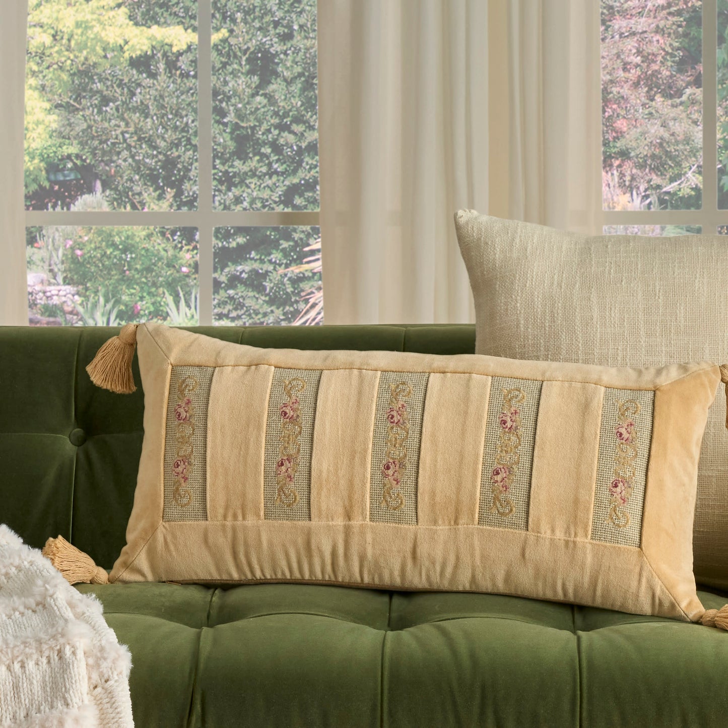 Beige rectangular decorative pillow with vertical floral-embroidered stripes and tassels on a green tufted sofa.
