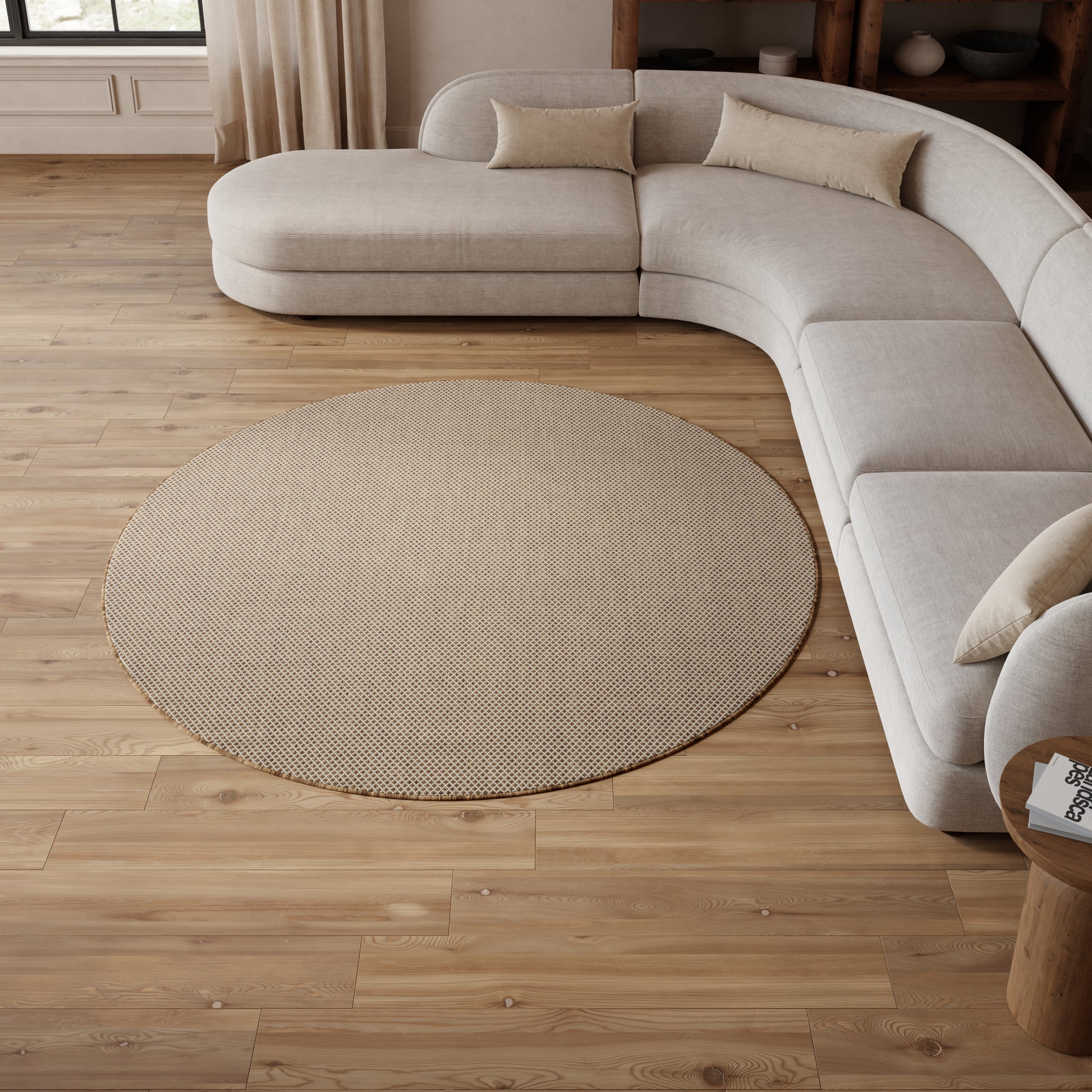 Light beige curved sectional sofa with plush cushions and a round textured beige rug on warm wooden flooring.