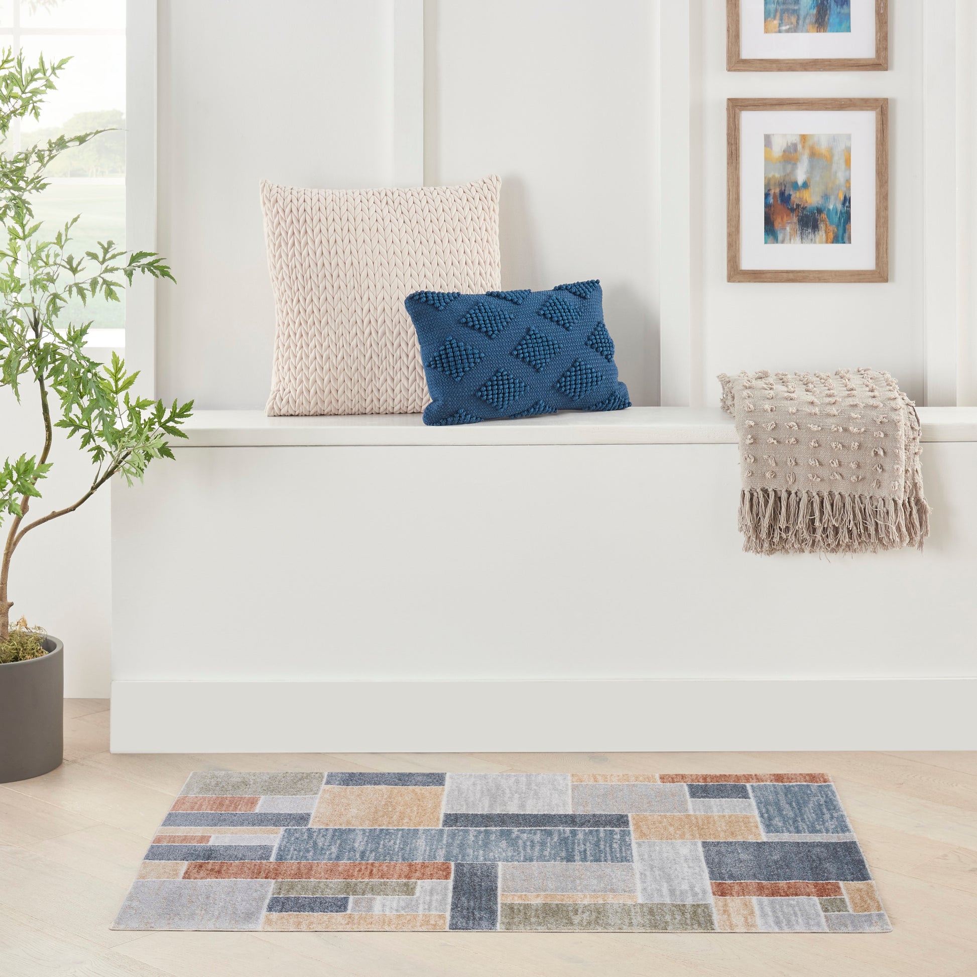 Contemporary 3' x 5' color-block rug as the main subject, with a blue diamond-knit pillow and beige knitted cushion on a white bench, and a fringed beige throw nearby.