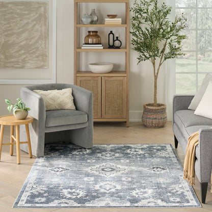 Cozy living room with grey upholstered chairs, light wood shelving, blue patterned area rug, potted tree, and wicker basket.
