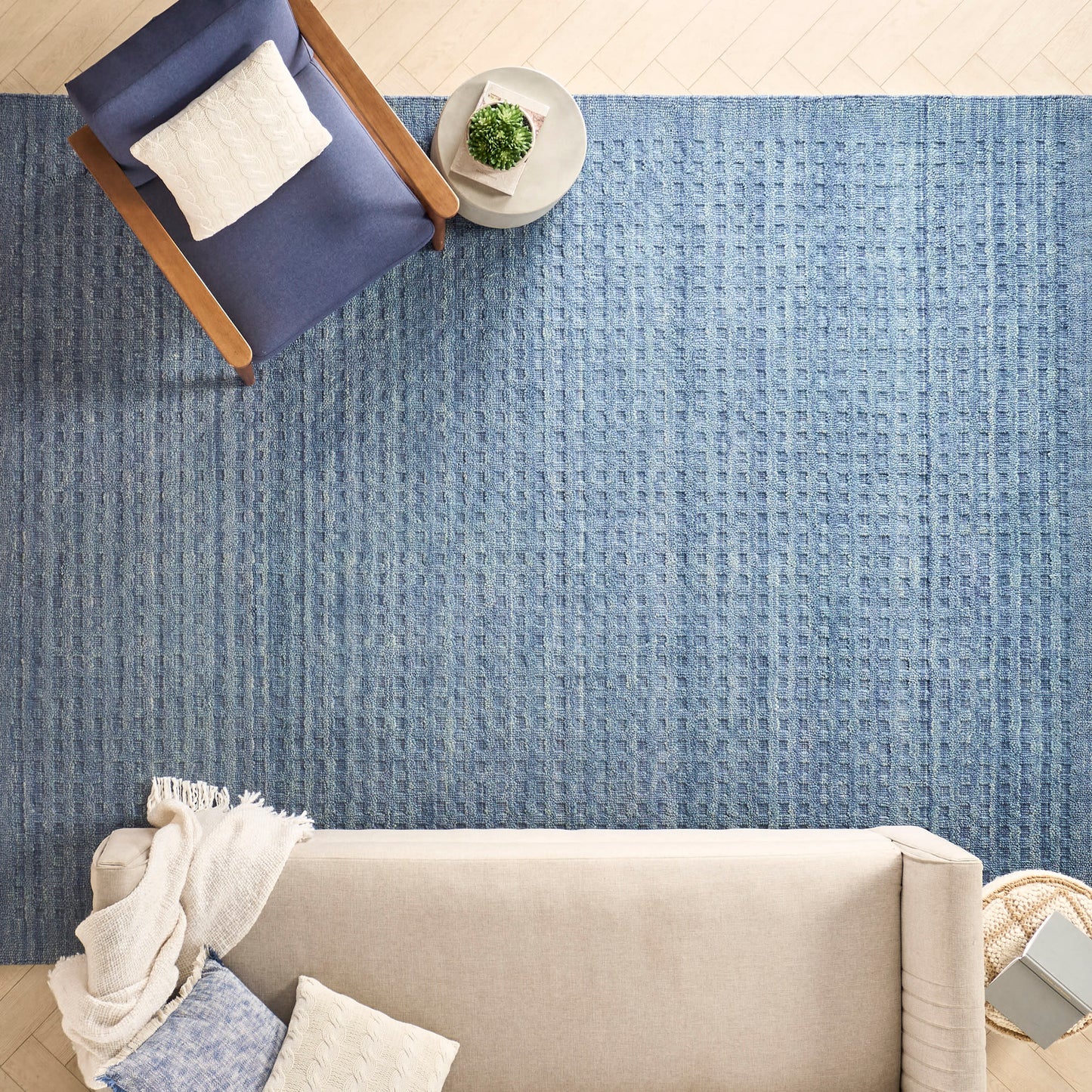 Overhead view: blue textured rug, beige sofa with cream throw, navy wood-armchair with white cushion, small round table with plant