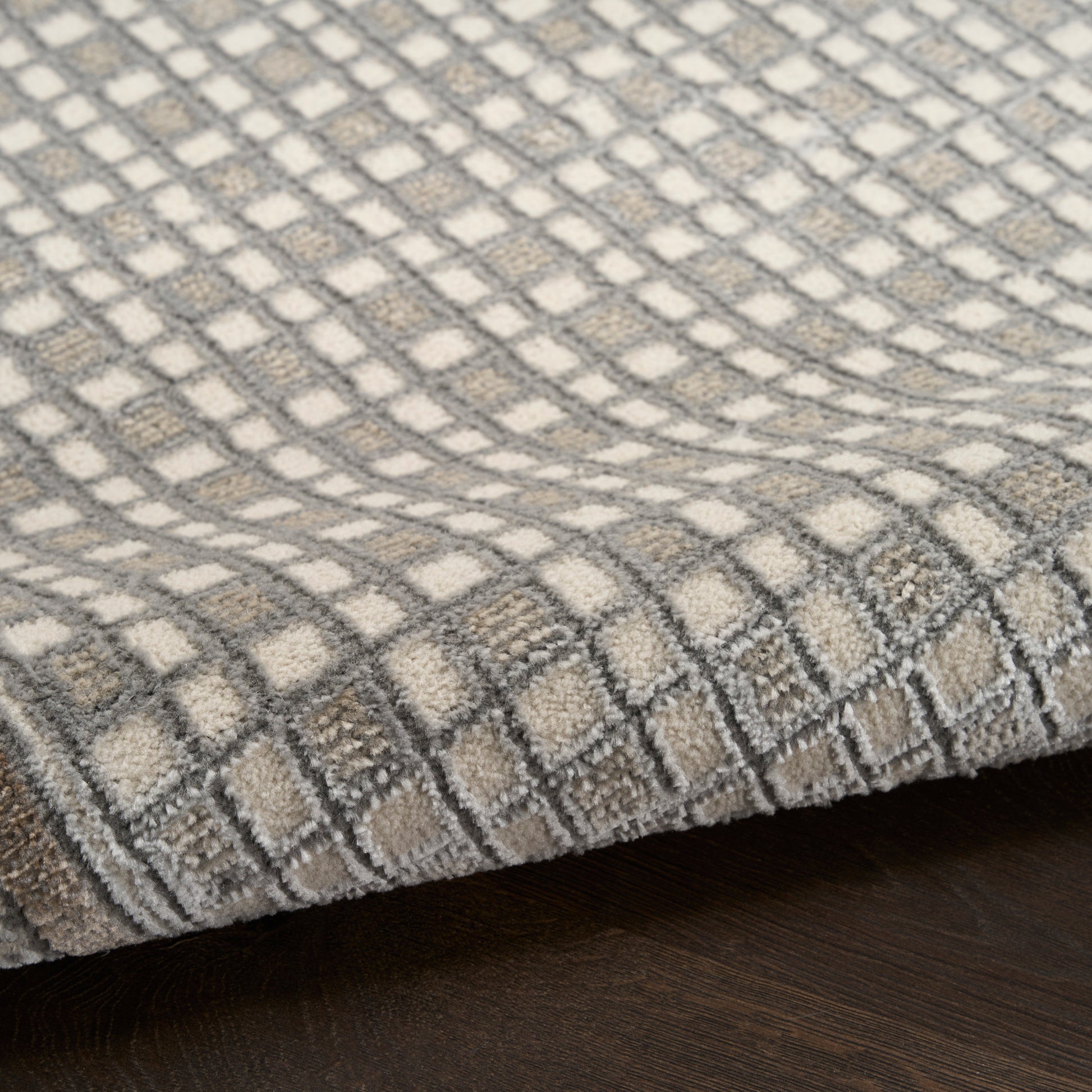 Beige and gray checkered woven rug with a soft loop pile, folded edge on dark wood floor.