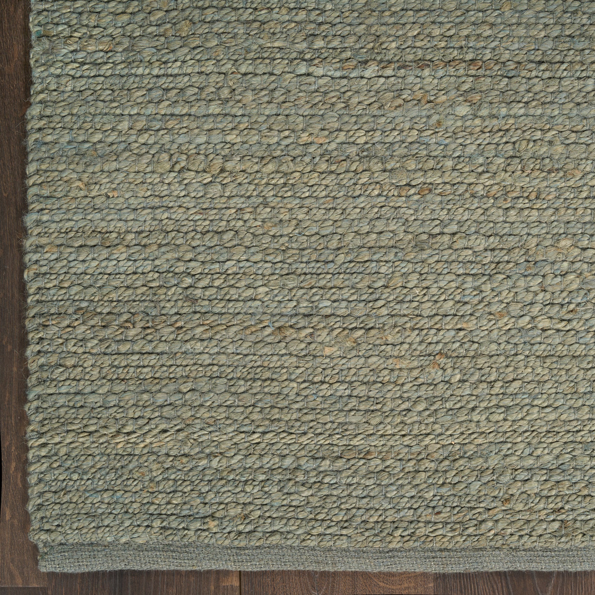Blue-gray looped-pile rug with beige flecks and dark brown border on wood floor.