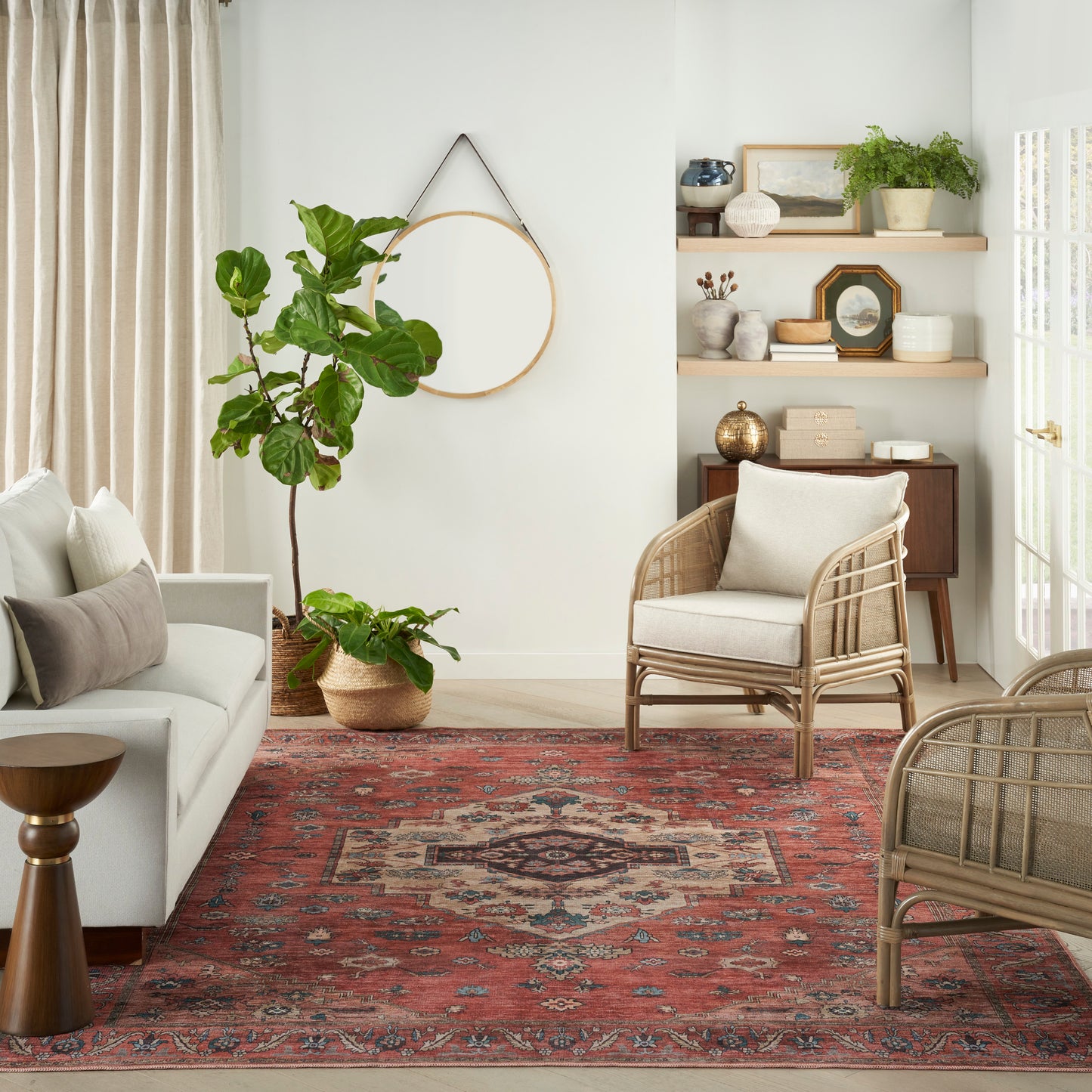 Boho living room with a red geometric rug, white sofa, woven rattan chairs, tall potted plant, round wall mirror, and wooden shelves.