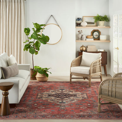 Boho living room with a red geometric rug, white sofa, woven rattan chairs, tall potted plant, round wall mirror, and wooden shelves.