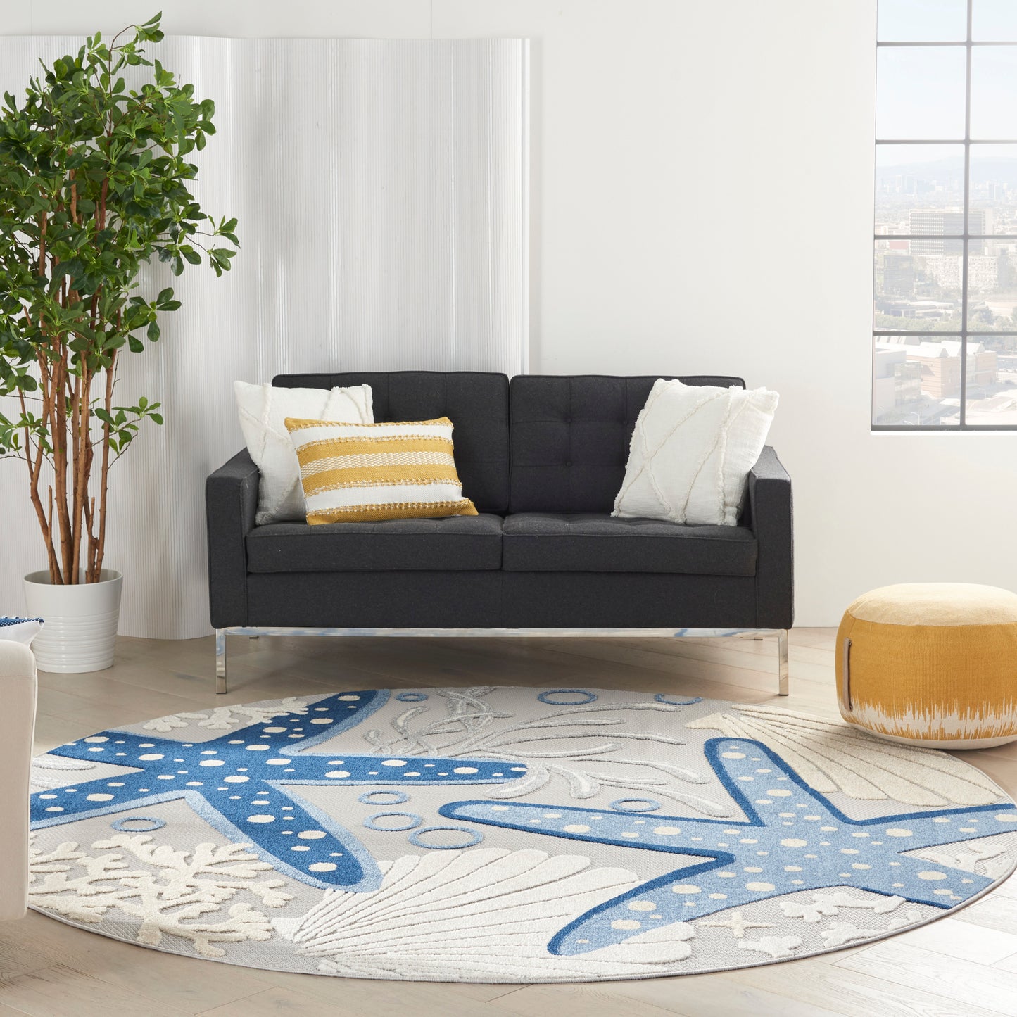 Round coastal rug (8' diameter) with a blue starfish pattern; in a living room with a dark gray fabric sofa on metal legs, white/yellow cushions, a potted plant, and a yellow pouf.