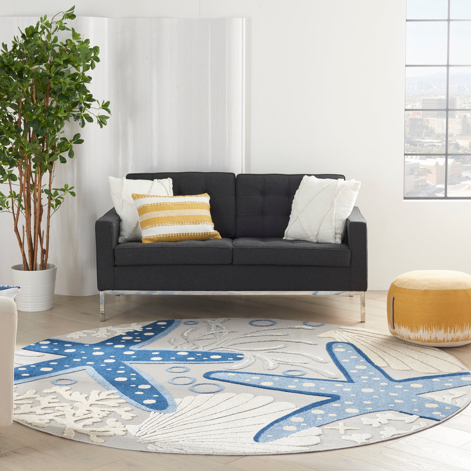 Round coastal rug (8' diameter) with a blue starfish pattern; in a living room with a dark gray fabric sofa on metal legs, white/yellow cushions, a potted plant, and a yellow pouf.