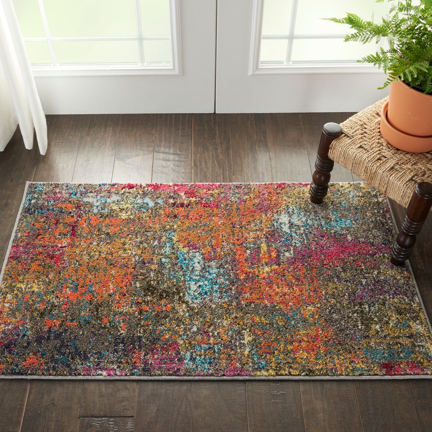 Colorful abstract shag rug in orange, pink, blue and purple hues on dark wood floor beside a woven bench with a fern.