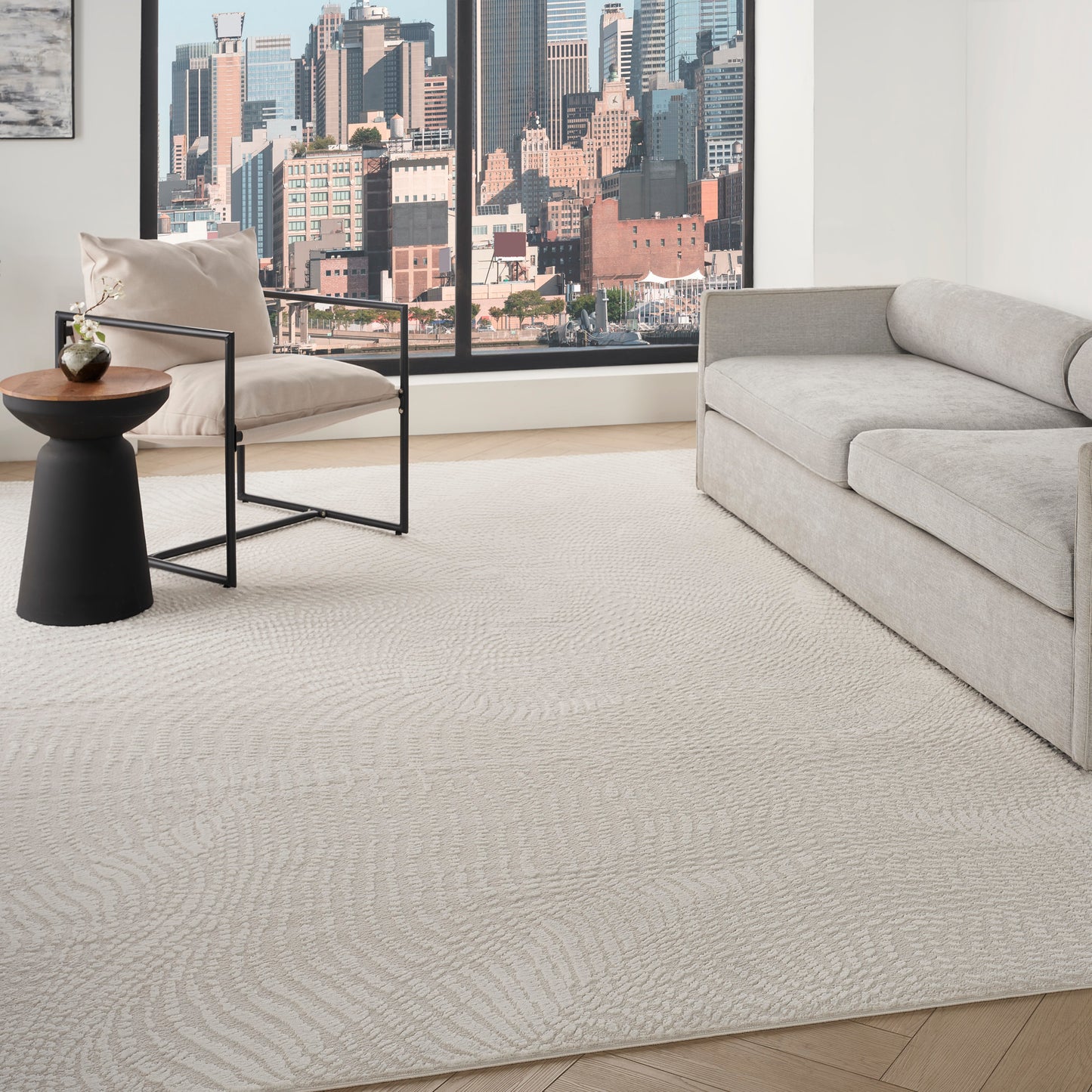 Beige area rug with subtle circular pattern in a modern living room with gray sofa, black metal chair, and round side table.