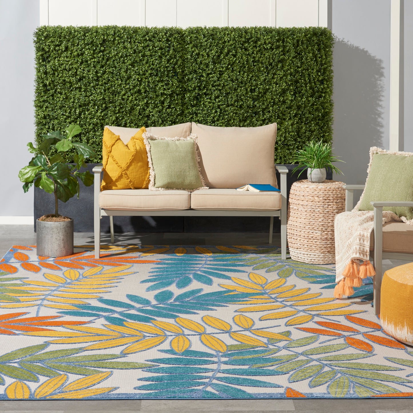 Outdoor lounge with beige sofa, green hedge backdrop, colorful pillows, woven side table, and a vibrant leaf-pattern rug.