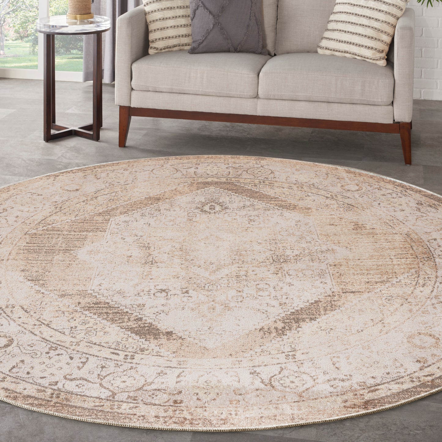 Round beige distressed oriental rug with faded ivory medallion, placed in a living room with a light sofa and wooden legs.