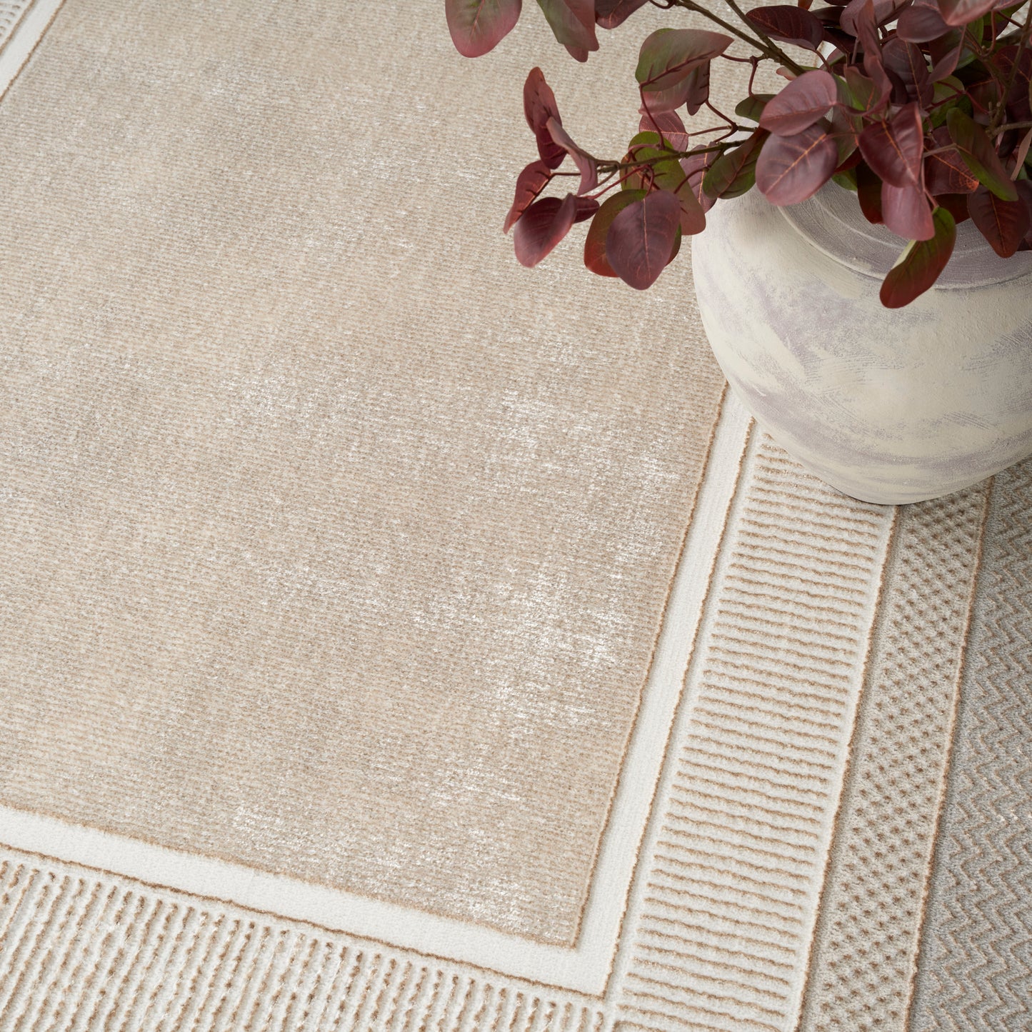 Beige textured area rug with layered borders; burgundy-leaf plant in a white pot rests on it.
