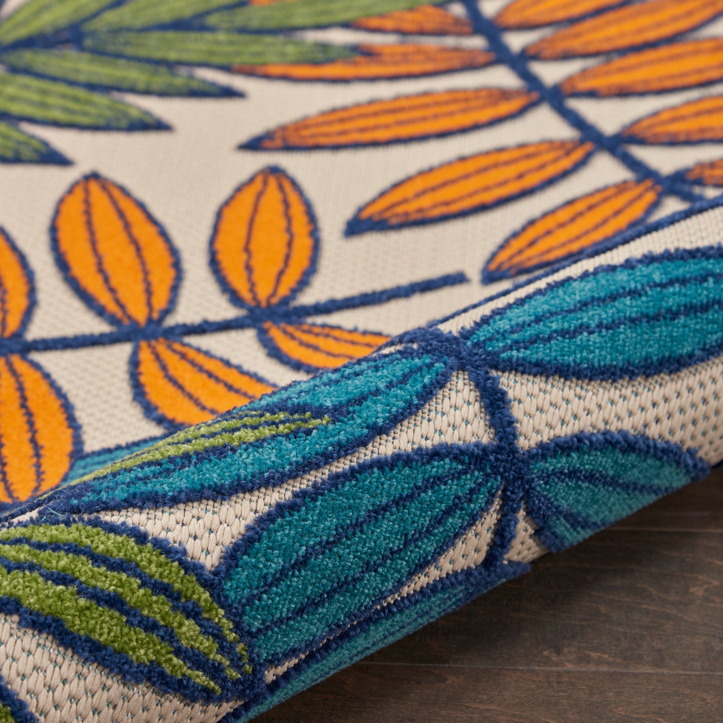 Close-up of a woven rug with teal leaves, orange seed shapes and olive accents on tan; textured fabric, folded edge.