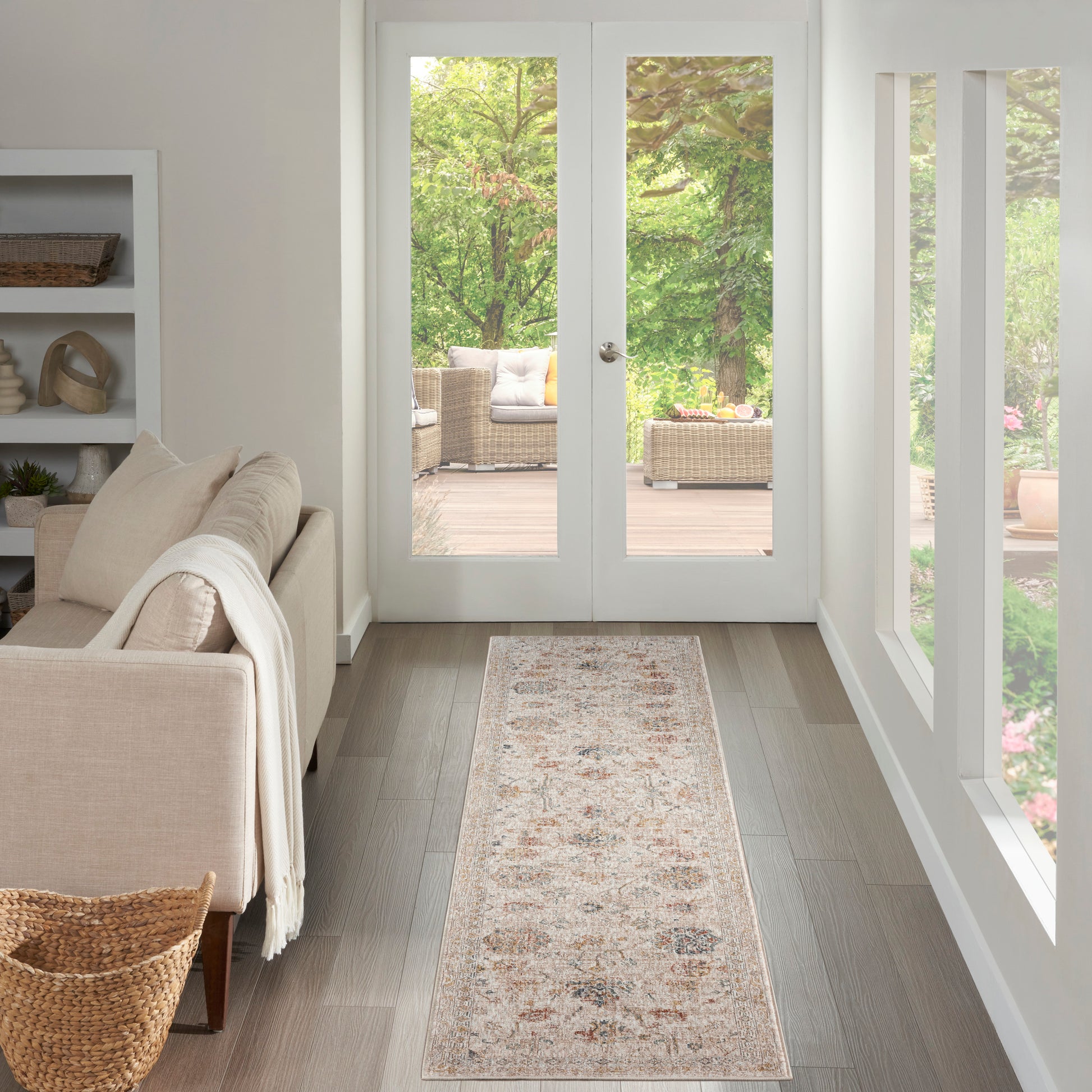 Beige fabric sofa with cream throw, light wood floor, long neutral rug runner, white walls, glass doors to outdoor wicker seating.