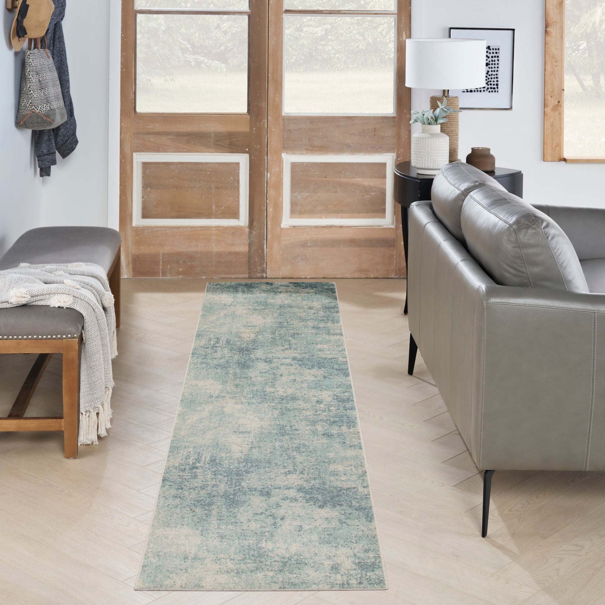 Blue-green distressed 8' runner rug on pale wood floor in a modern living room entry; gray leather sofa, bench, and wooden doors.