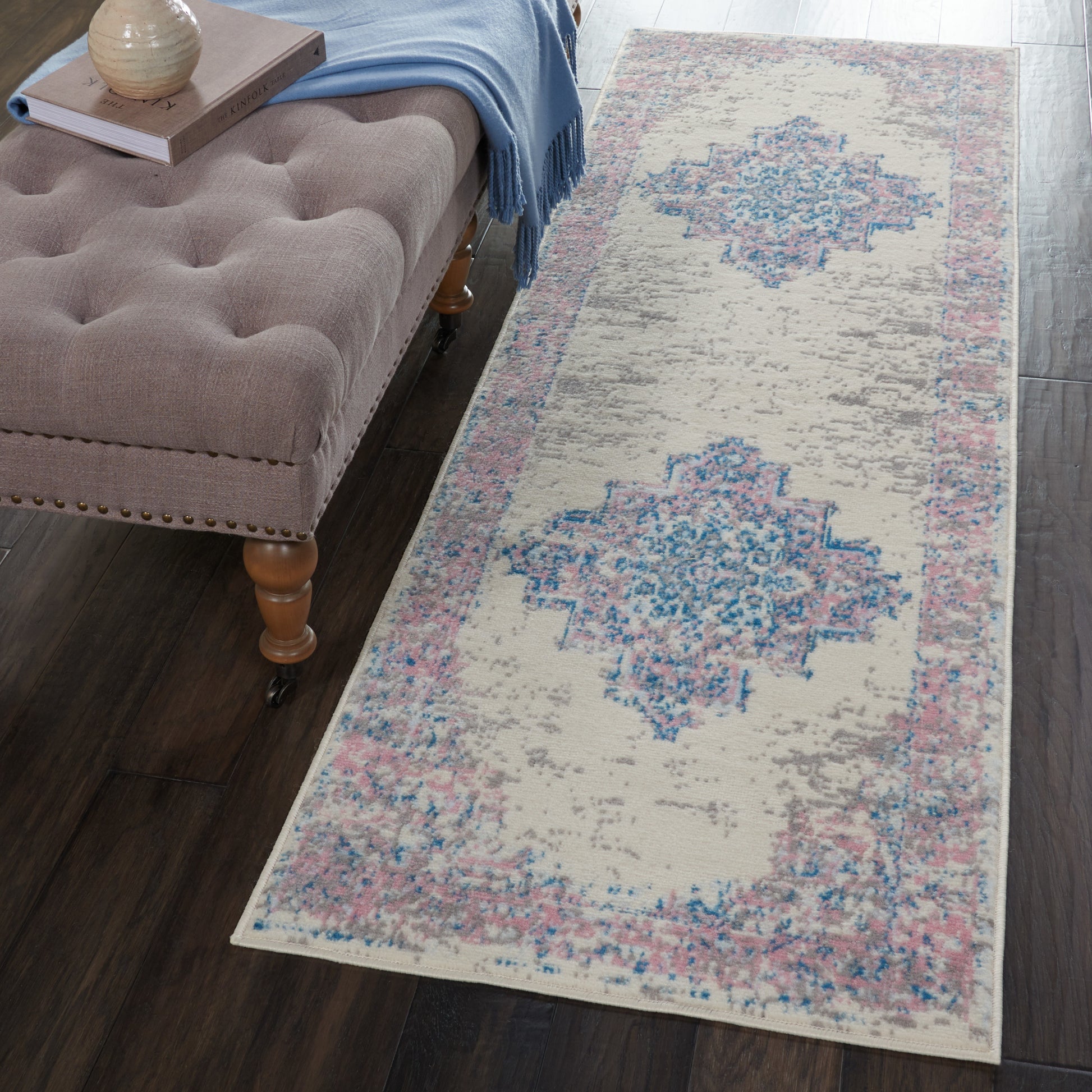 Distressed beige runner rug with blue & pink geometric medallions beside a tufted beige ottoman on dark wood floor.