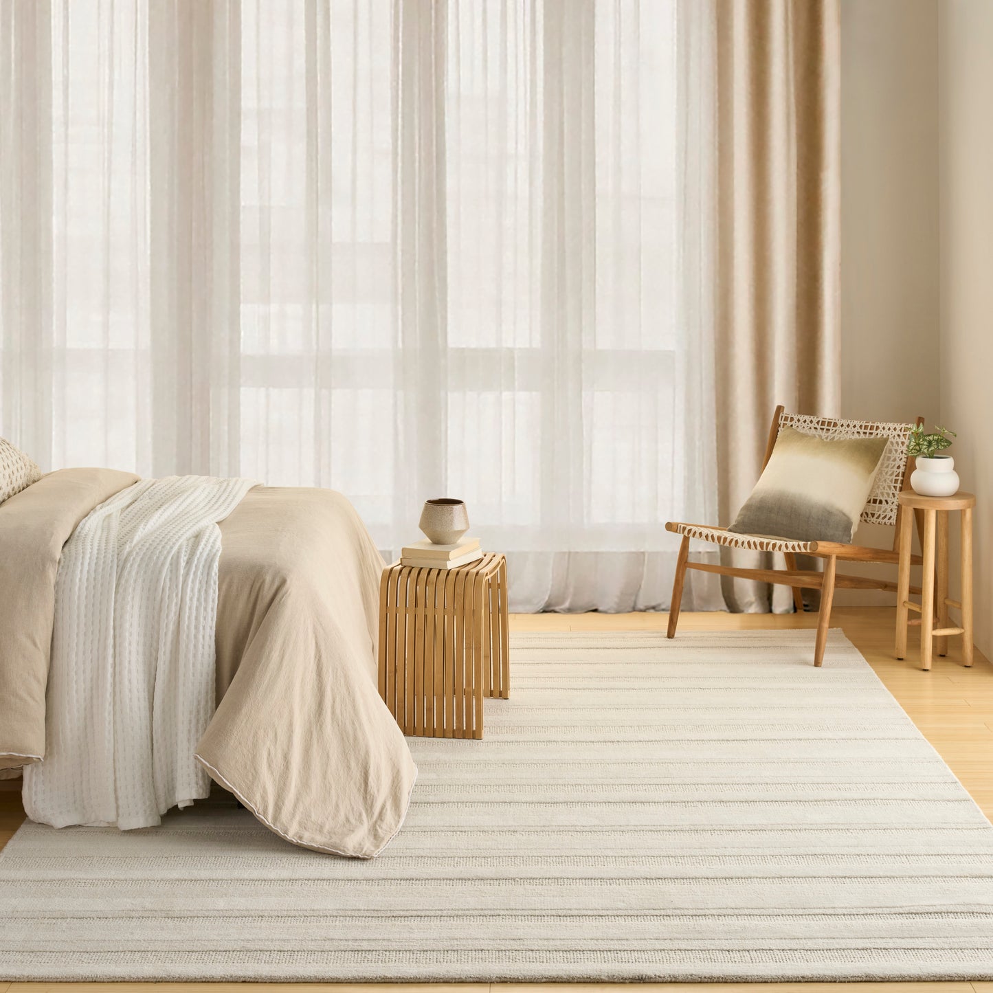 Neutral-toned bedroom with beige bedding, sheer curtains, striped rug, wooden chair, and plant on a side table.