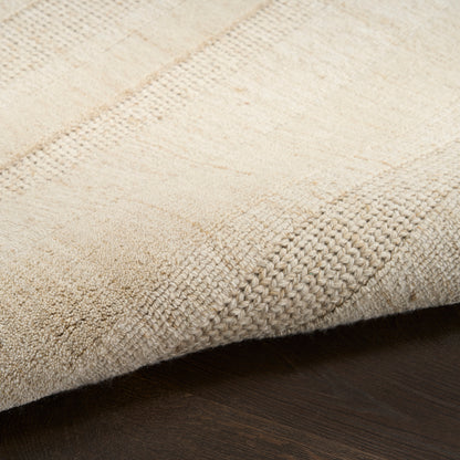 Beige woven area rug with subtle patchwork stripes & soft loop pile, edge folded on dark wood floor.