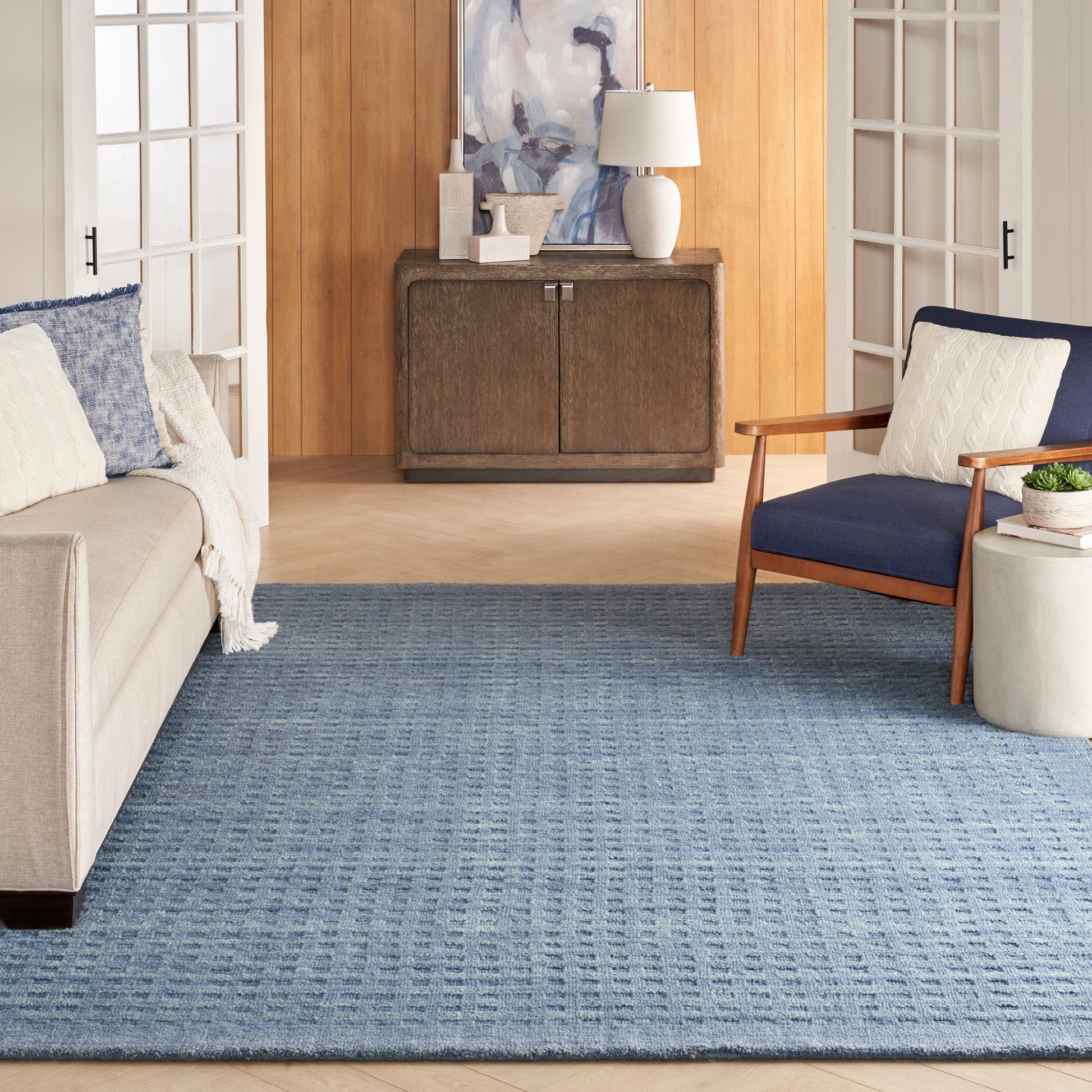 Blue textured area rug in a cozy living room with beige sofa, navy chair, and rustic wood cabinet.