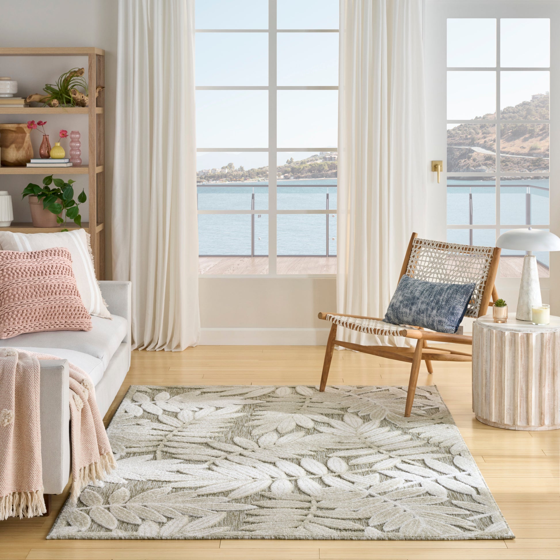 Coastal living room with gray leaf-pattern rug, white sofa with pink throw, woven armchair, light wood floor and sea-view windows.