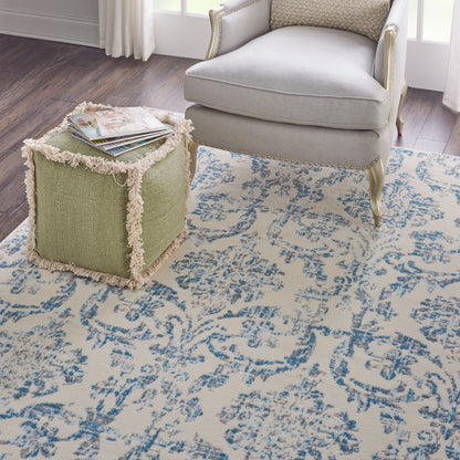 Blue damask rug on the floor with a light gray upholstered armchair and a green cube ottoman with fringe trim and magazines on top.