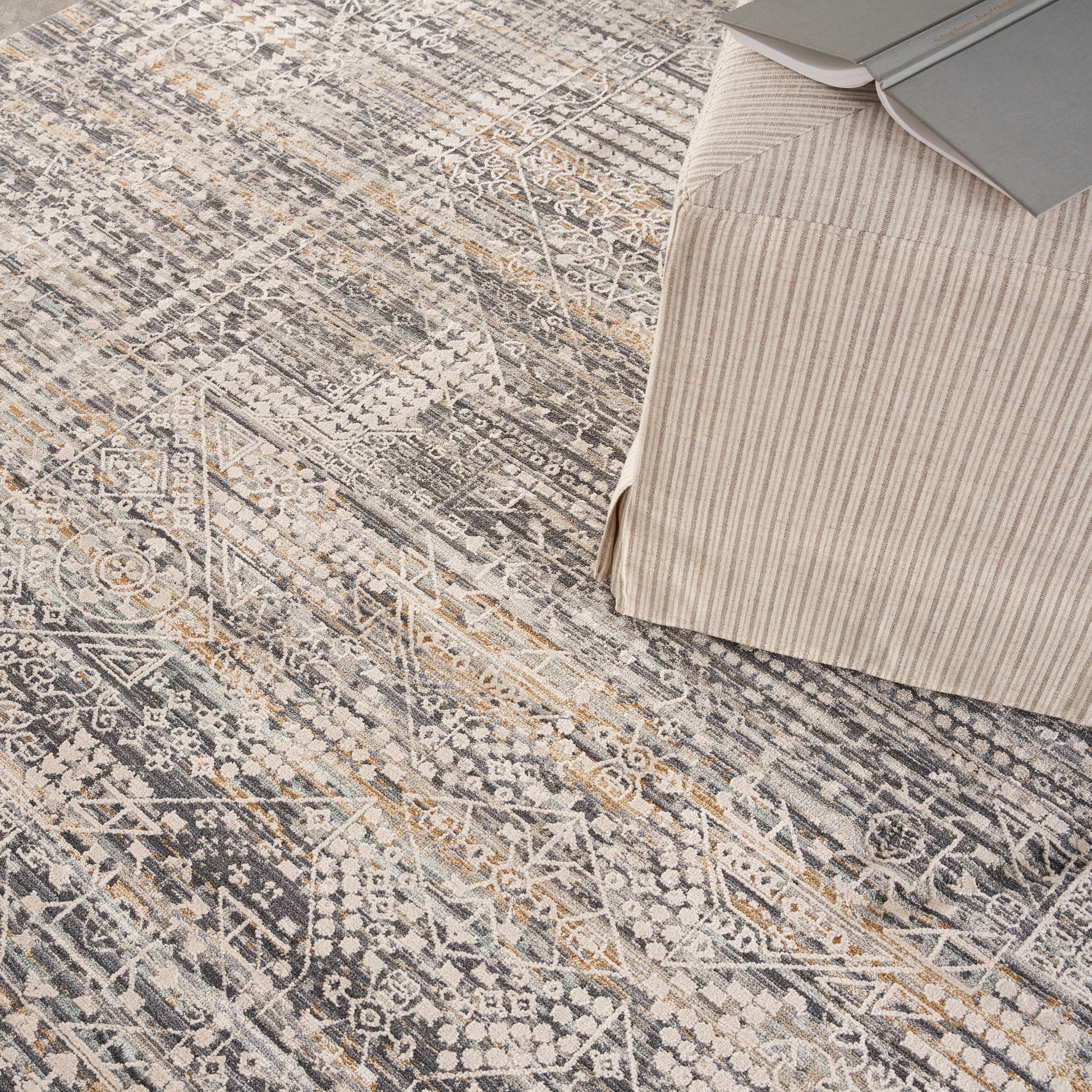Geometric patterned area rug in gray, beige and ivory beside a beige striped ottoman with books on top.