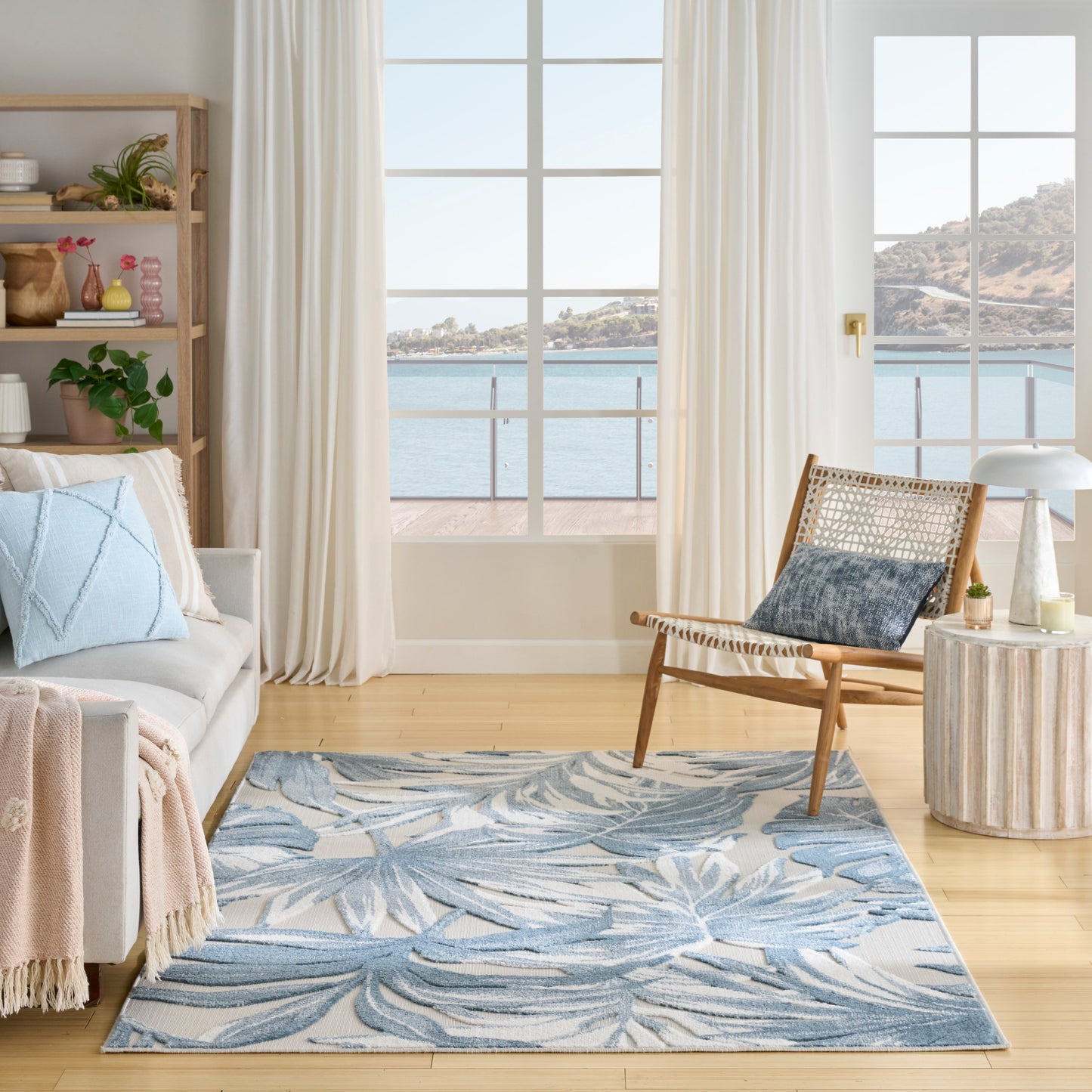 Bright coastal living room, blue palm-leaf rug, light gray sofa, woven wood lounge chair, white curtains, sea-view windows.