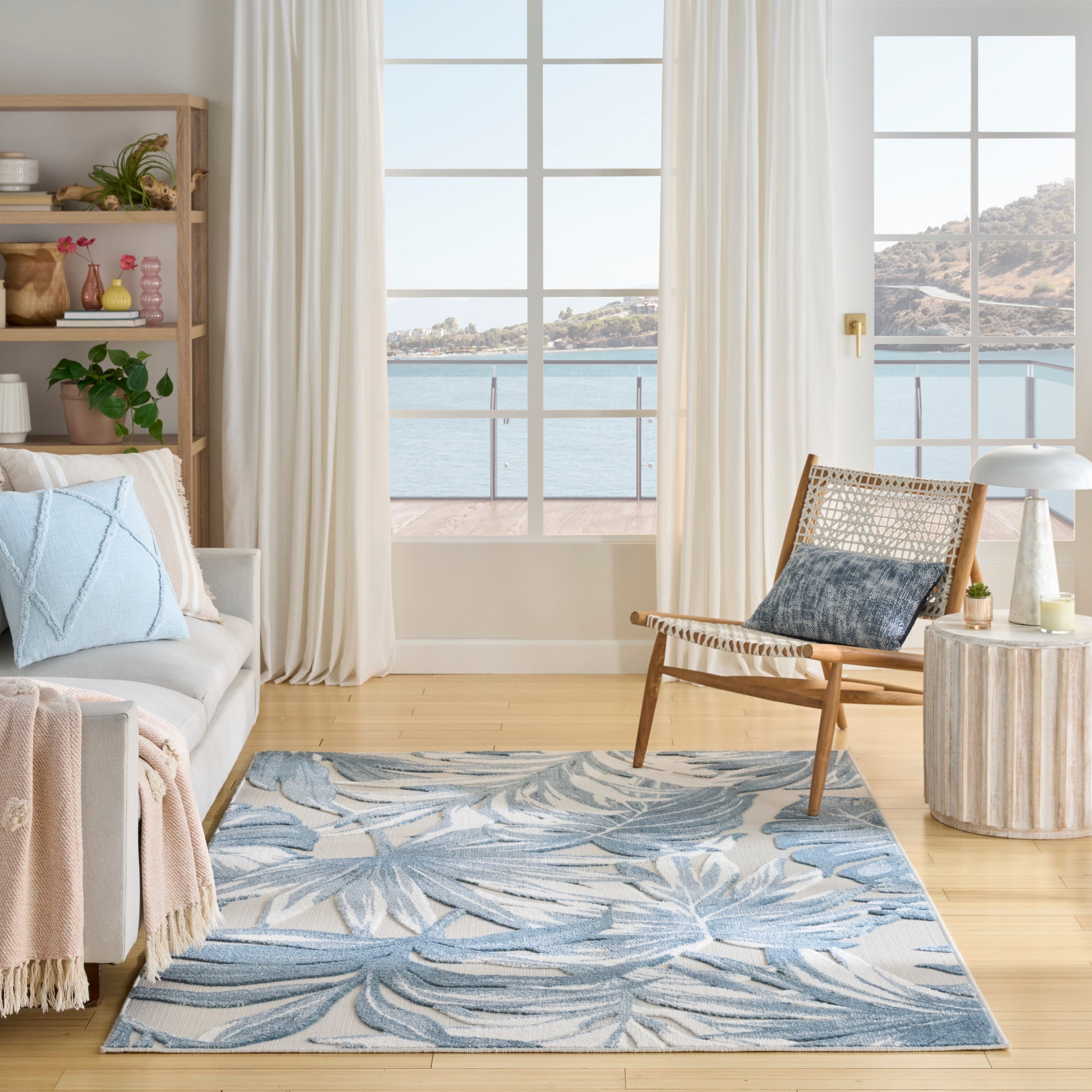 Bright coastal living room, blue palm-leaf rug, light gray sofa, woven wood lounge chair, white curtains, sea-view windows.