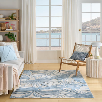 Bright coastal living room, blue palm-leaf rug, light gray sofa, woven wood lounge chair, white curtains, sea-view windows.
