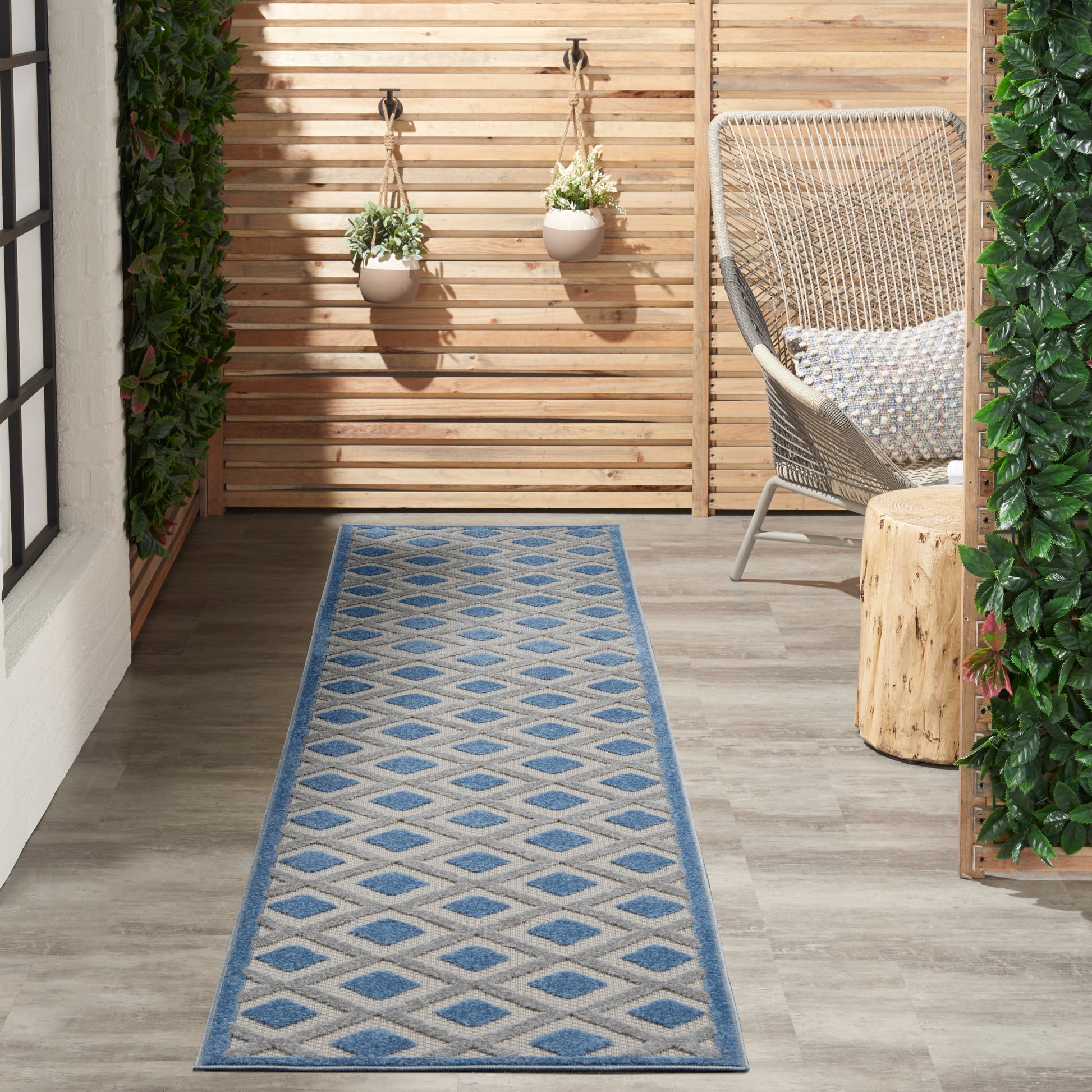 Outdoor runner rug in blue & beige diamond pattern on a wooden deck, with hanging planters and a woven chair.