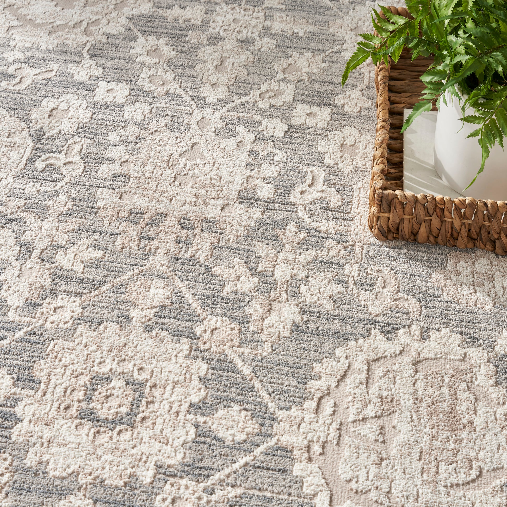 Gray and ivory patterned rug with a damask-like motif; wicker basket with a green fern at the edge.