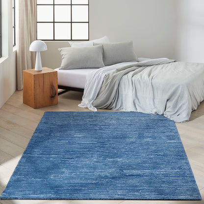 Blue rectangular area rug with subtle texture on light wood floor beside bed with grey duvet.