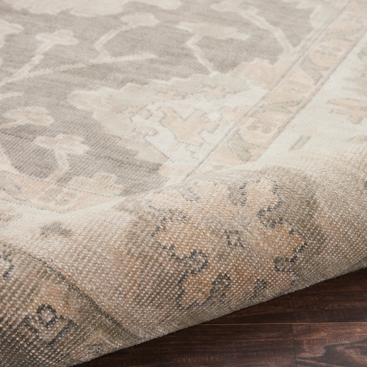 Neutral woven rectangular area rug in beige and gray with a floral medallion pattern, folded corner on dark wood floor.