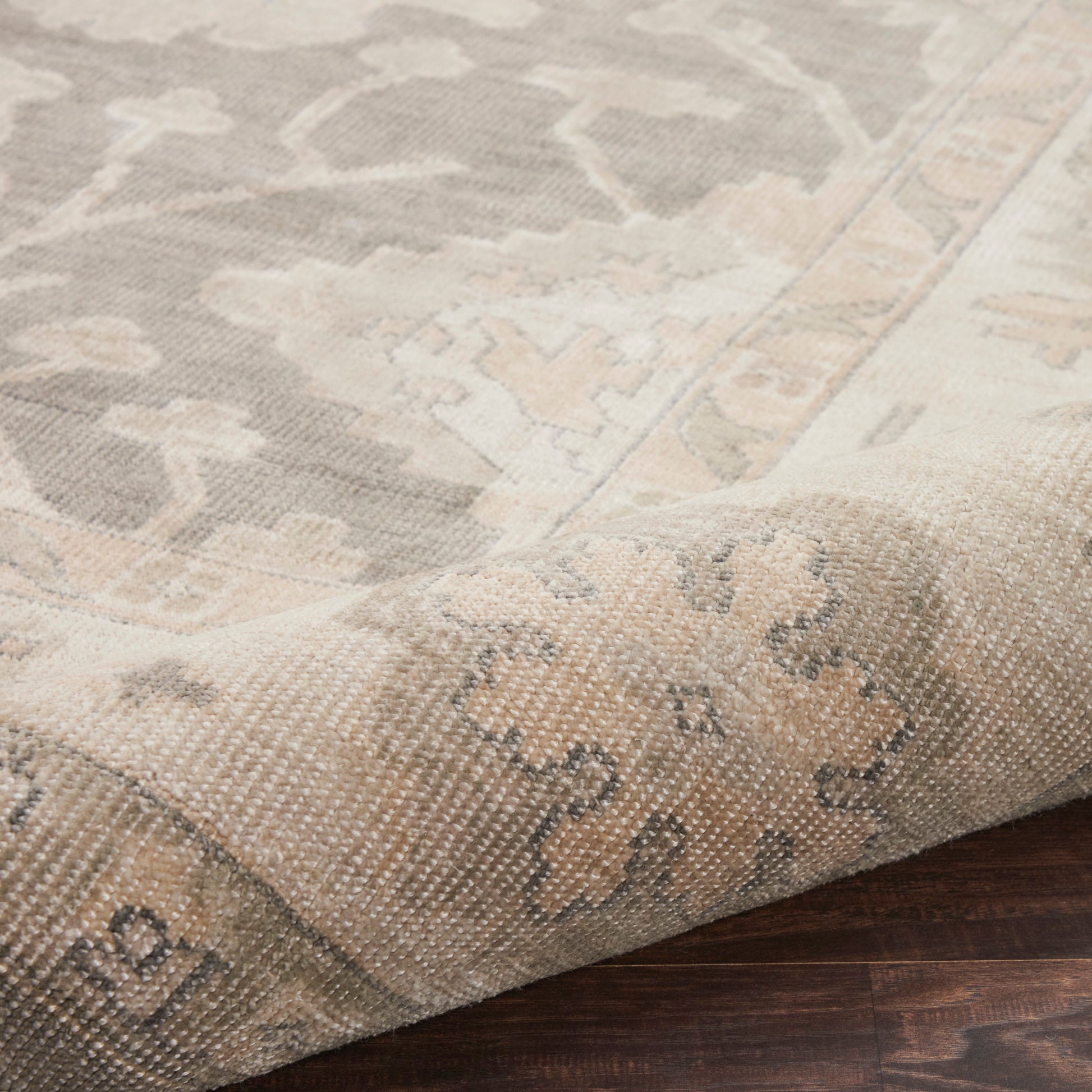 Neutral woven rectangular area rug in beige and gray with a floral medallion pattern, folded corner on dark wood floor.