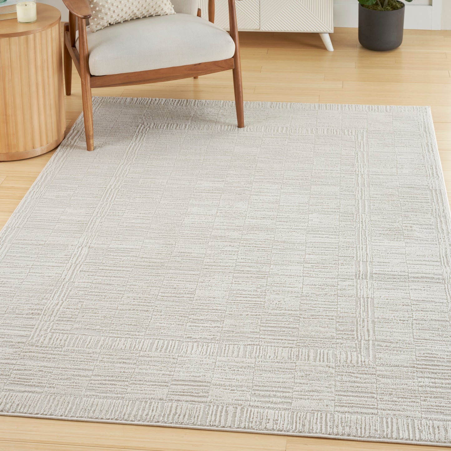 Neutral beige woven area rug with a subtle grid border on a light wood floor in a cozy living room with a wooden chair.