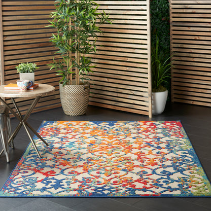 Rectangular woven rug with bright multicolor pattern (blue, orange, red, yellow, green) on dark floor beside potted plants and wooden slats.