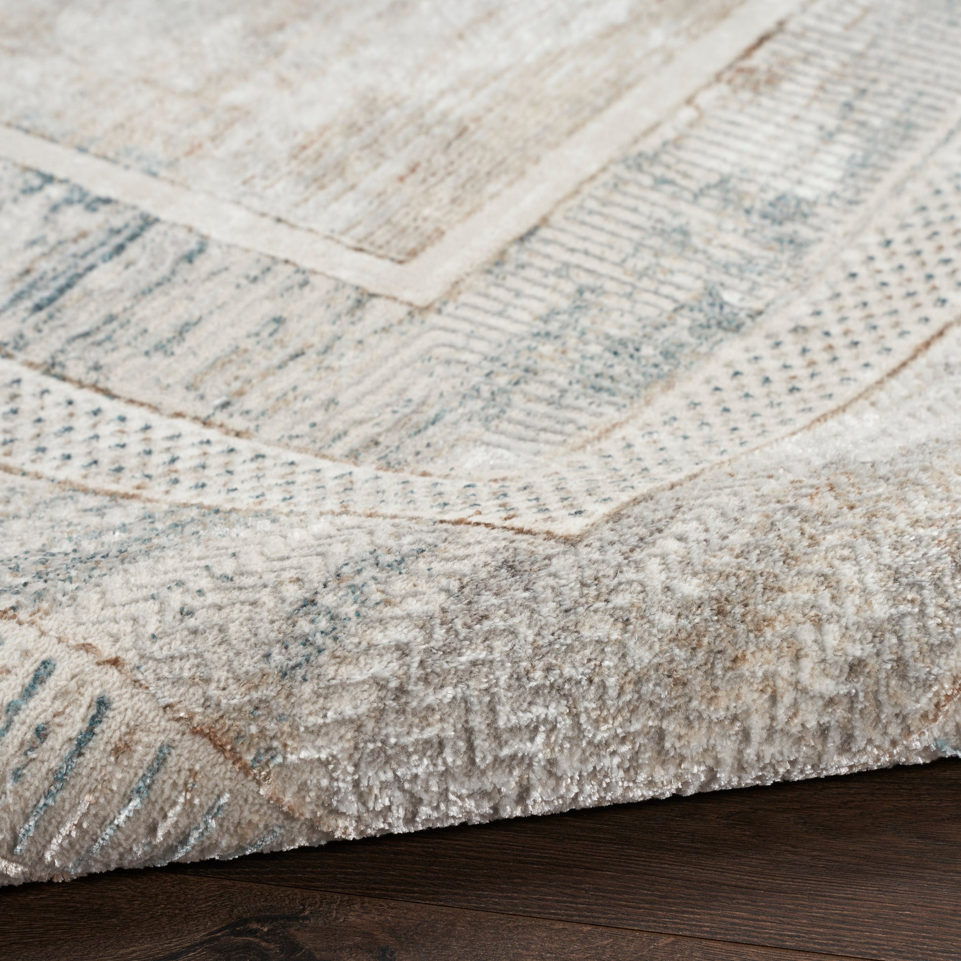 Close-up of a light beige-gray area rug with geometric panel pattern, folded corner, and soft textured pile on wood floor.