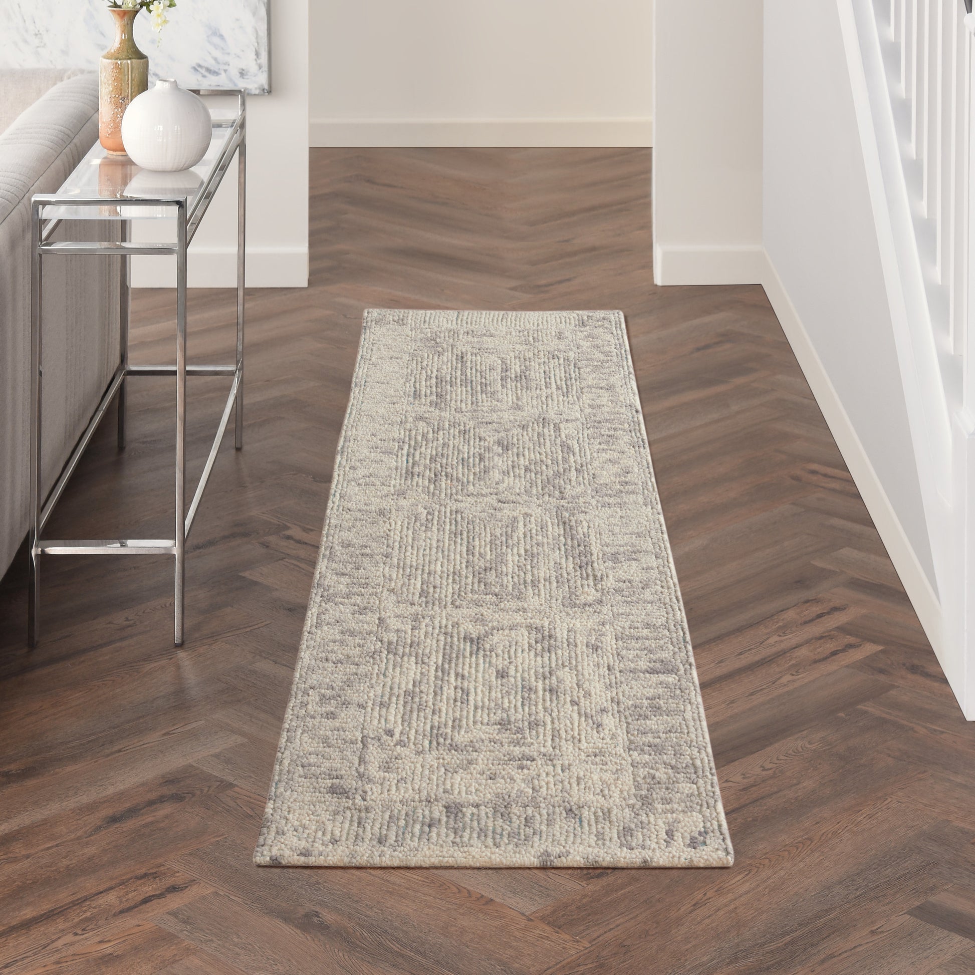 Neutral beige runner rug in a hallway with dark herringbone wood floor; glass-top, metal-framed console table on left.