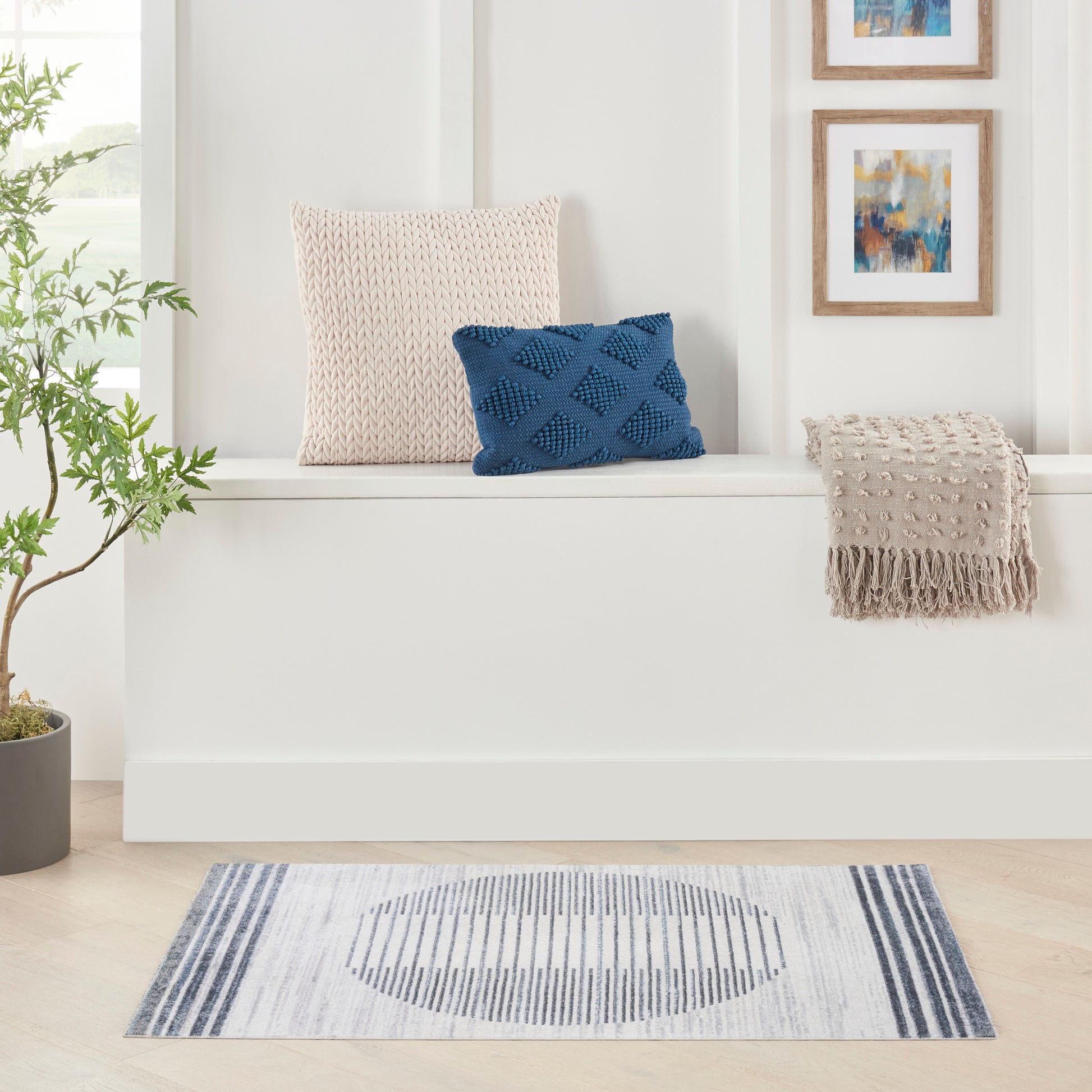 Blue striped mid-century modern rug 2' x 4' beneath a white built-in bench with beige knit cushion, blue lattice cushion, beige fringe throw, and a potted plant.