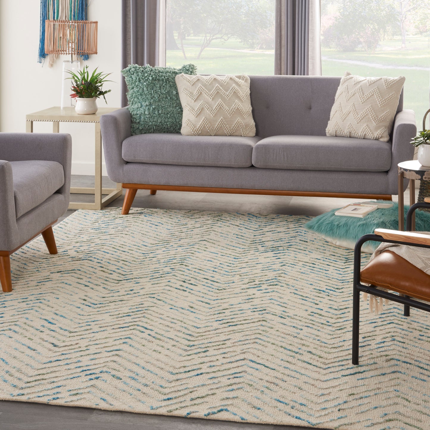 Grey fabric mid-century sofa with wooden legs, beige chevron pillows, teal throw, matching chair, and cream-blue zigzag rug.