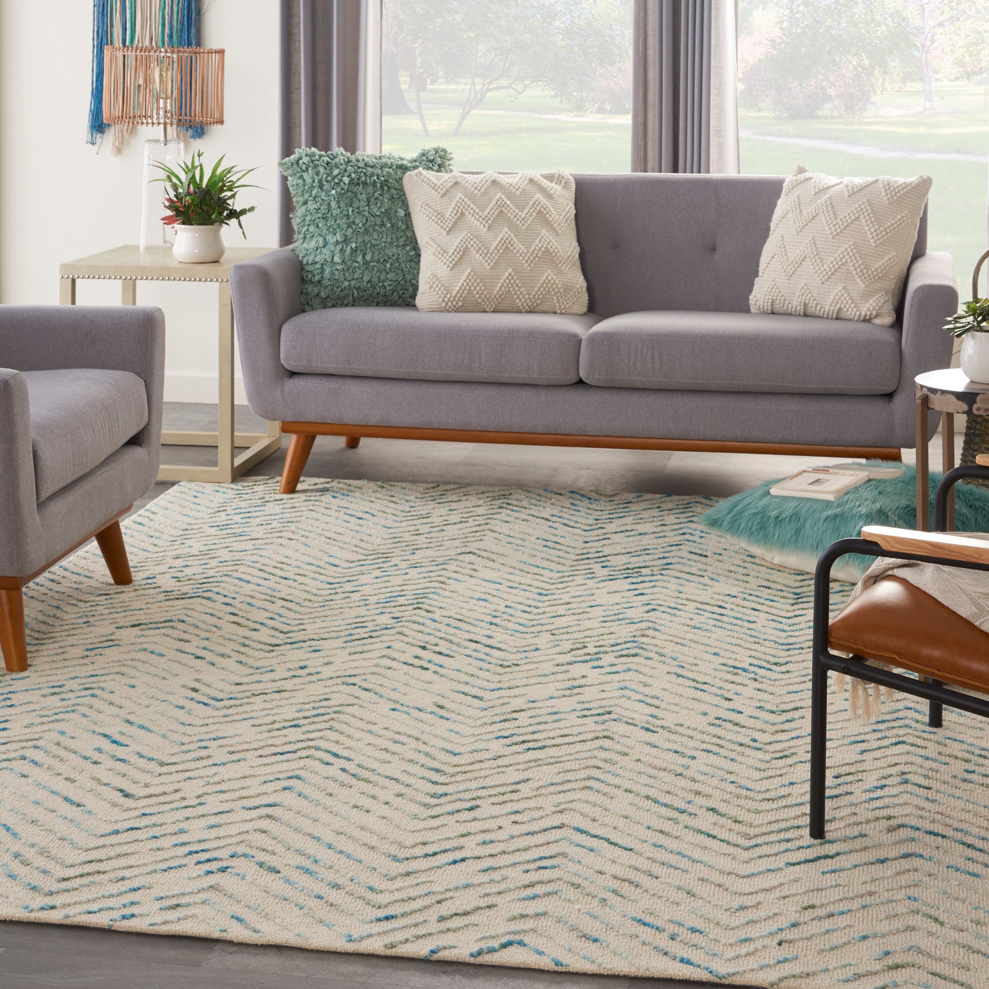 Grey fabric mid-century sofa with wooden legs, beige chevron pillows, teal throw, matching chair, and cream-blue zigzag rug.