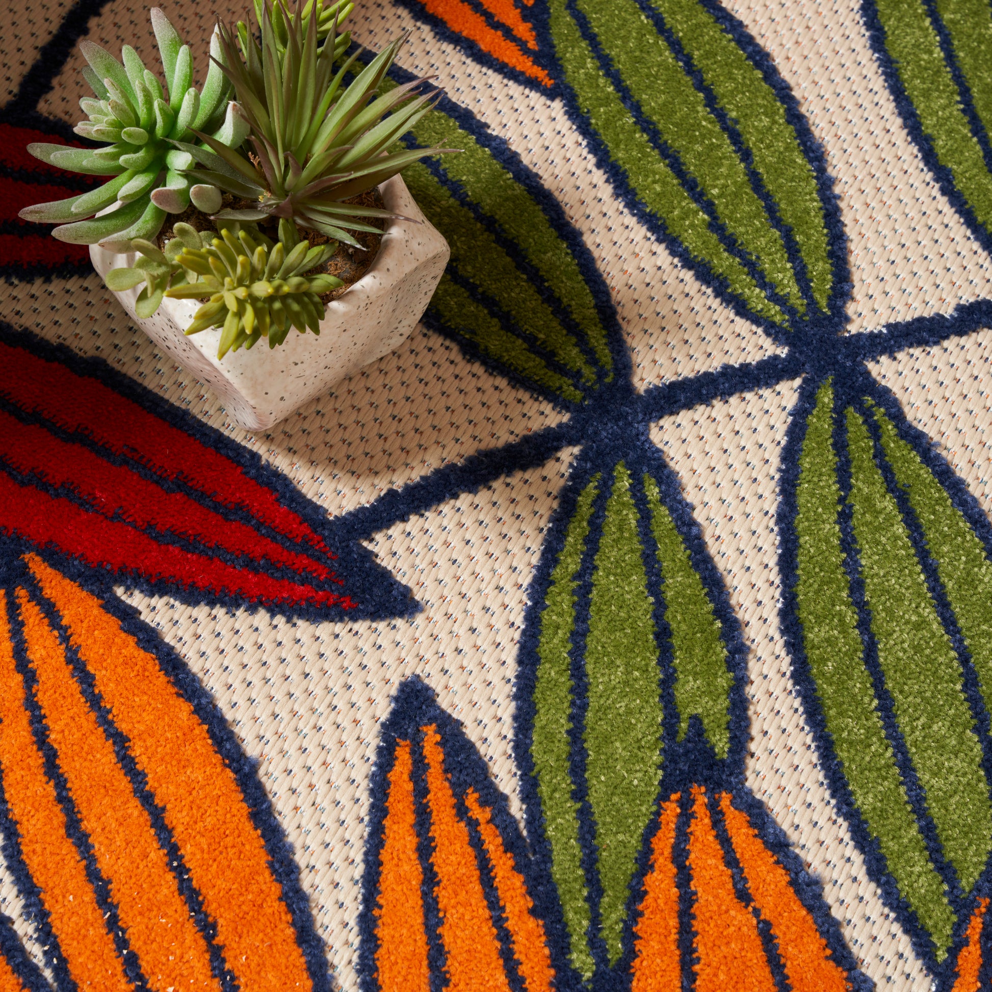 Beige woven rug featuring bold orange, green, and blue tropical leaves; small succulent pot rests on edge.
