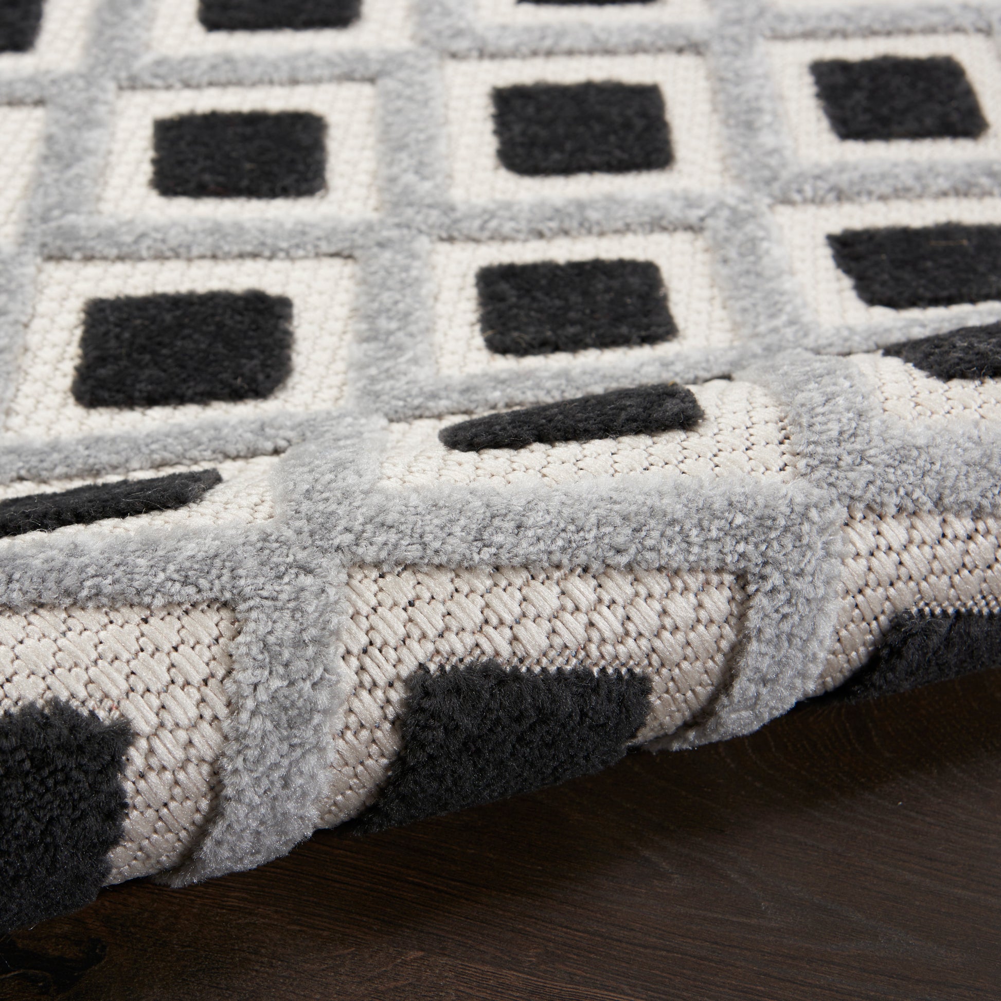 Soft gray rug with black square geometric pattern, plush pile and folded edge on dark wood floor.