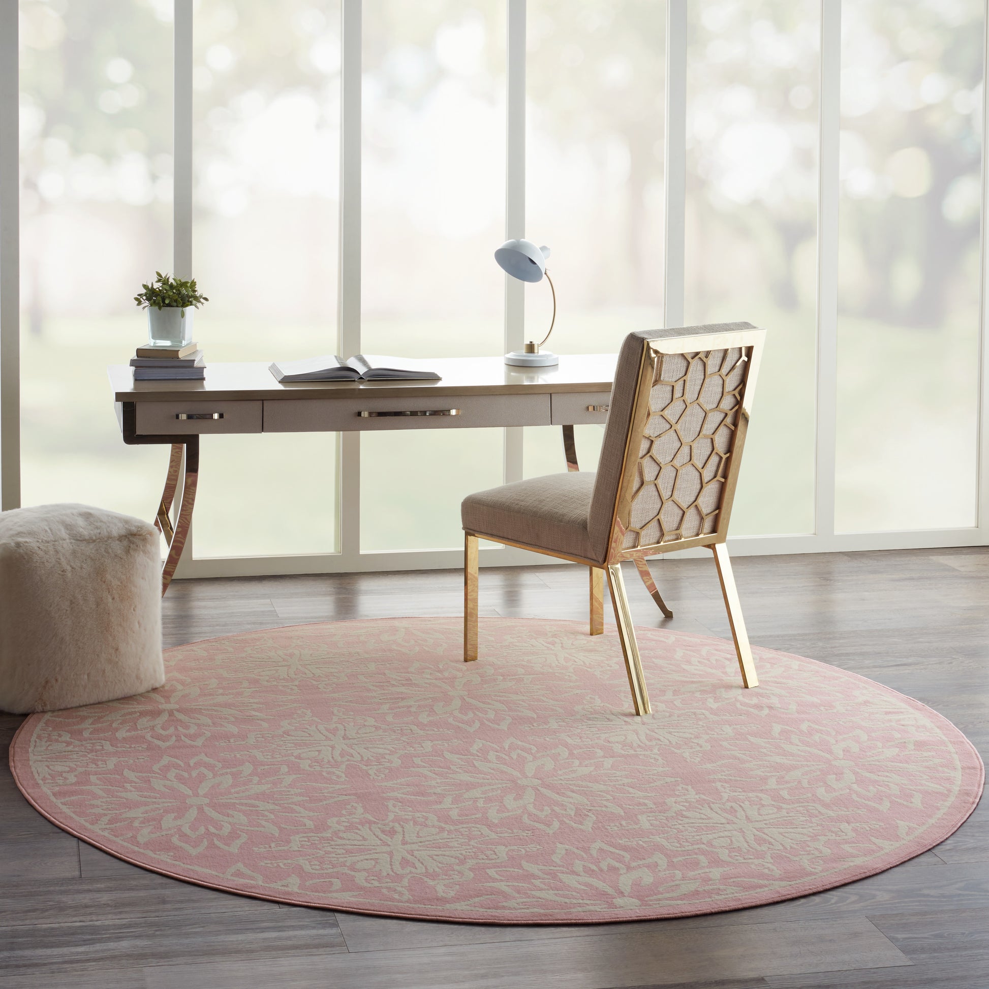 Pink round rug under light wood desk with beige honeycomb-pattern chair, plant, lamp and books by sunlit windows.