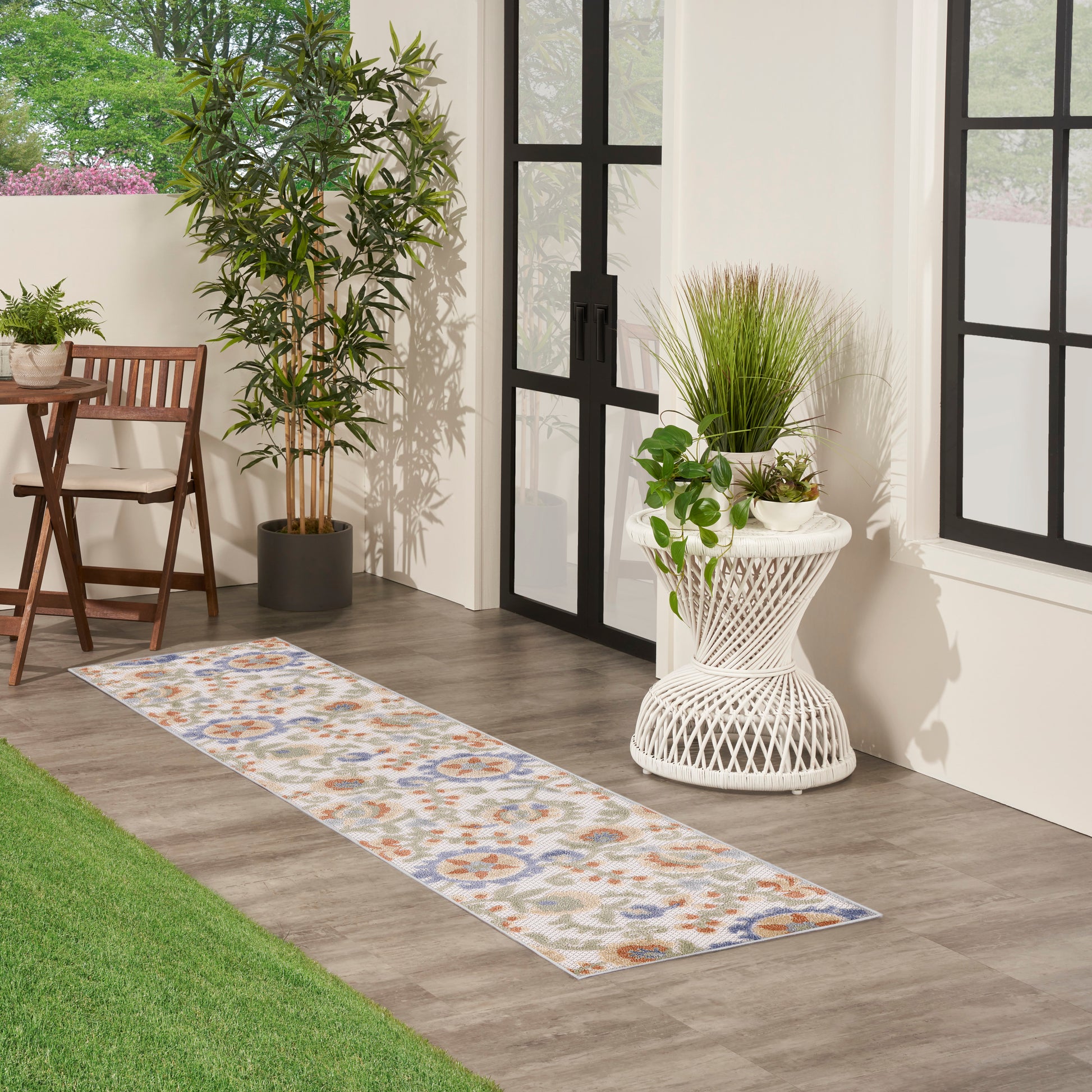 Beige runner rug with blue and orange floral medallions on gray wood floor beside potted greenery and white wicker planter.